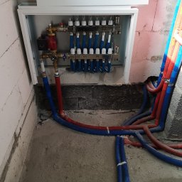 Aqua - Rozdzielacz ogrzewania podłogowego w białej skrzynce, z podłączonymi rurami w kolorze niebieskim i czerwonym, zamocowanymi do ściany z betonu komórkowego.