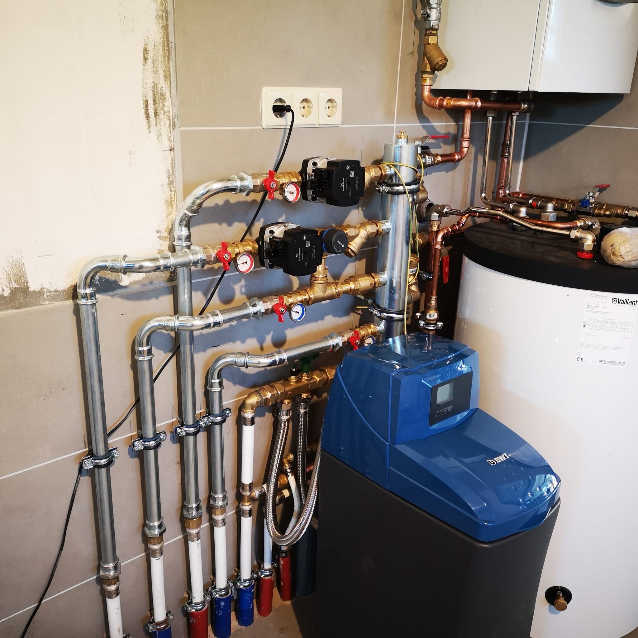 Złożona instalacja hydrauliczna z rurami miedzianymi i stalowymi, zaworami, pompami obiegowymi i zmiękczaczem wody BWT, zamontowana na tle płytek ceramicznych.