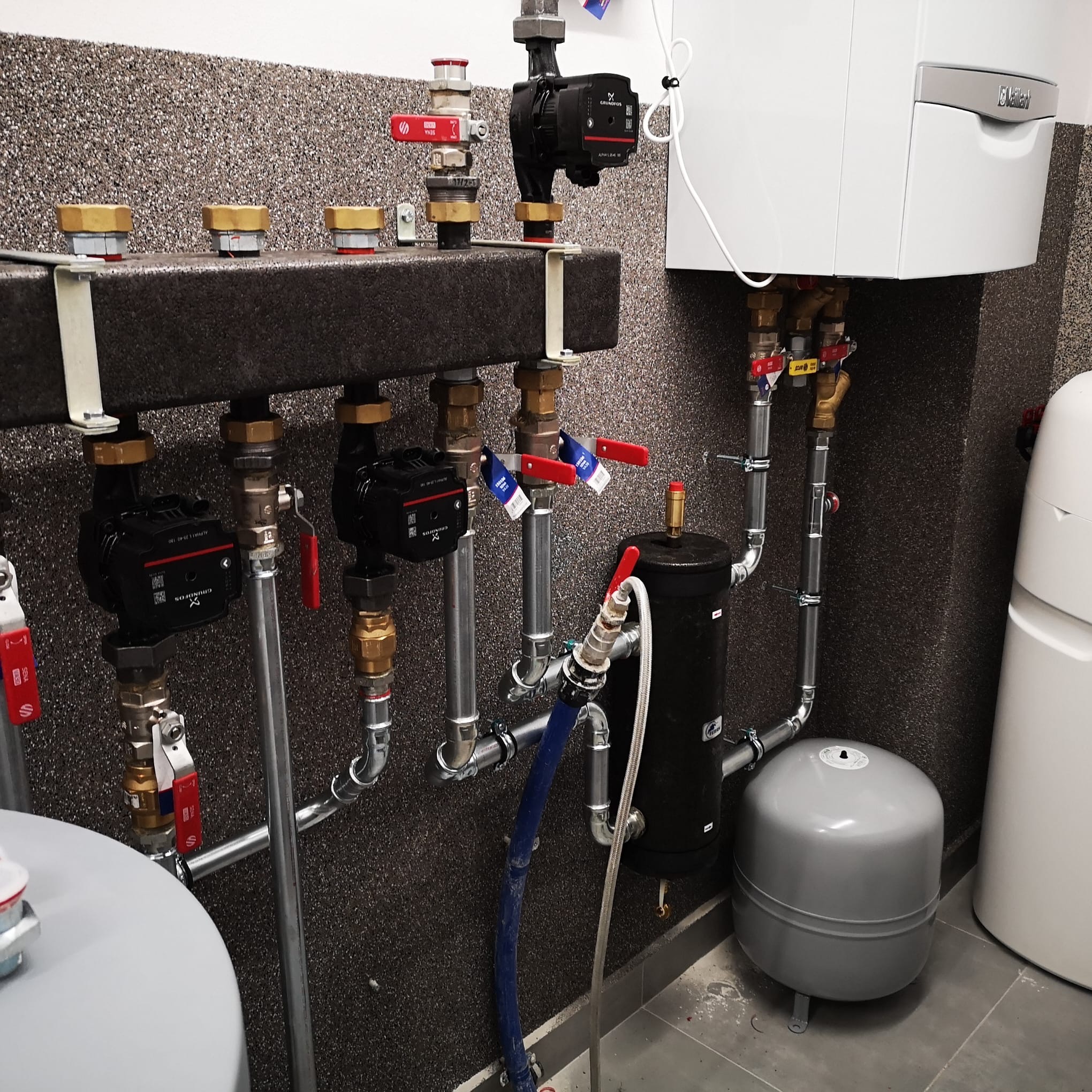 Nowoczesna kotłownia z piecem gazowym Vaillant, pompami obiegowymi Grundfos i zbiornikiem wyrównawczym. Instalacja z zaworami i rurami na tle ściany o fakturze baranka.