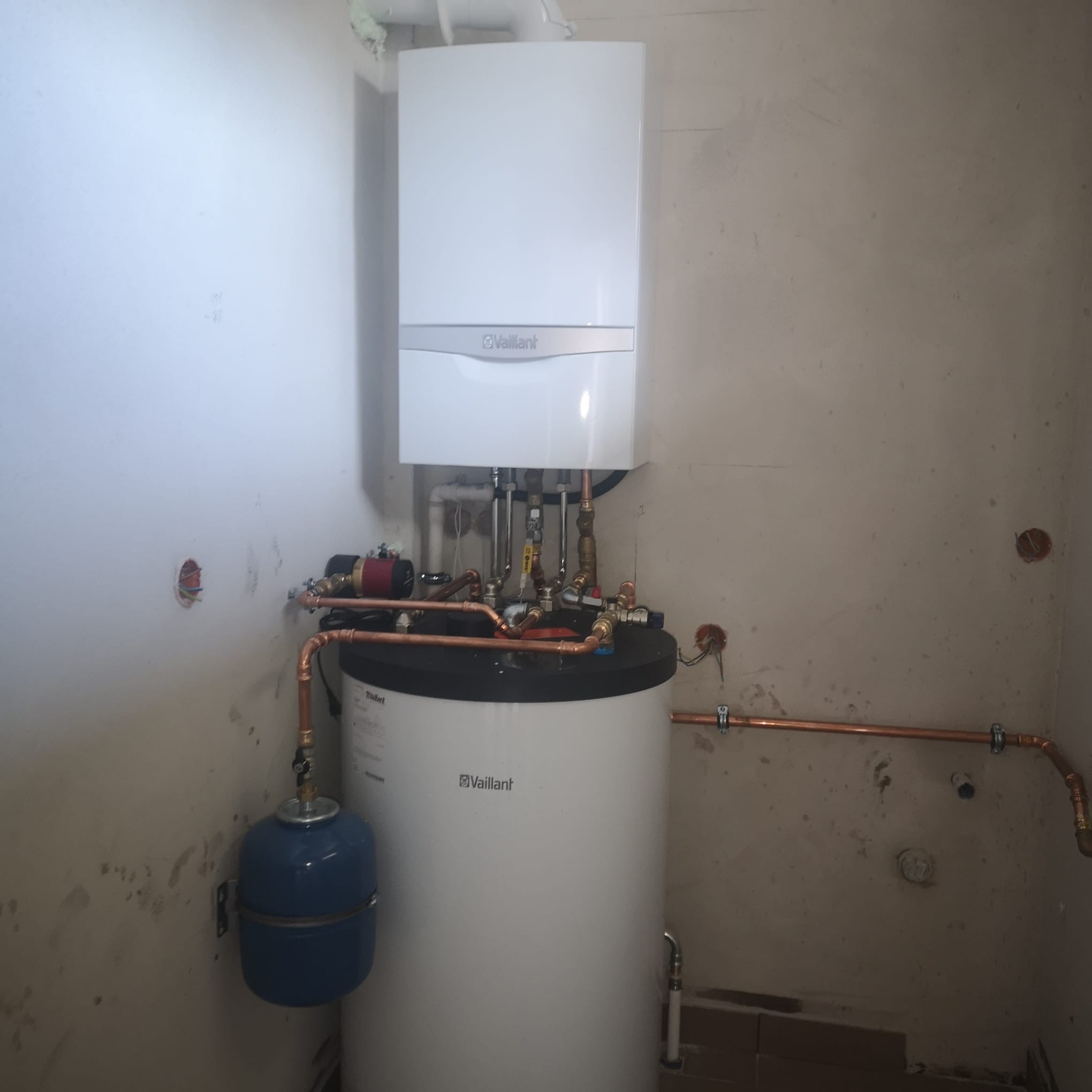 Biały piec gazowy Vaillant zamontowany nad zasobnikiem wody, z widocznymi miedzianymi rurami i elementami instalacji w surowym pomieszczeniu.