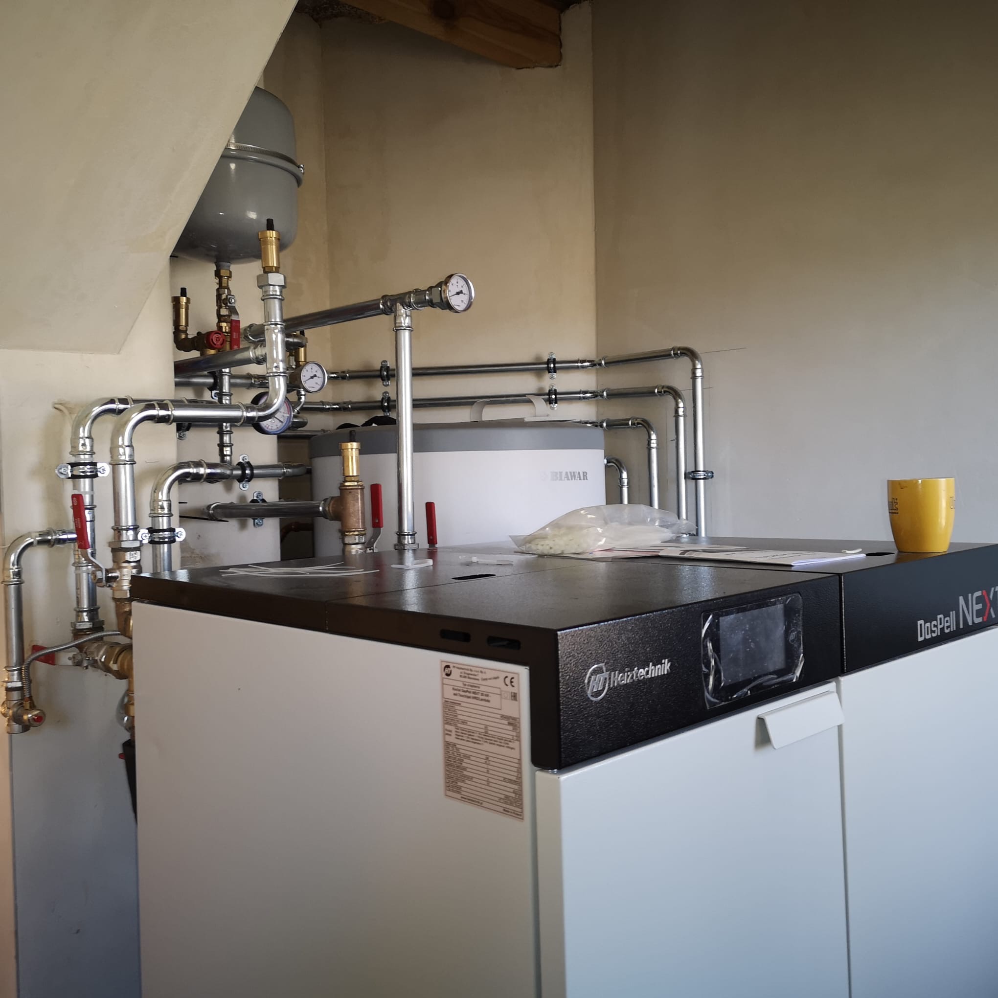 Nowoczesny piec DasPell NEXT z rozbudowaną instalacją hydrauliczną i zbiornikiem Biawar. Widoczny manometr i żółty kubek na piecu. Instalacja w jasnym pomieszczeniu.