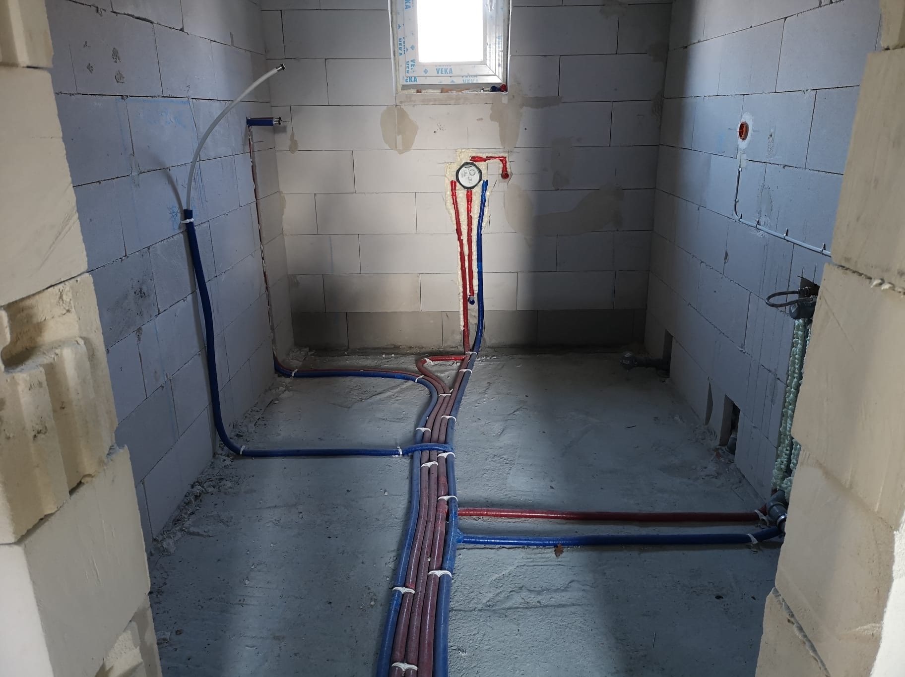 Instalacja wodna z rur PEX w surowym wnętrzu. Czerwone i niebieskie rury prowadzone wzdłuż ścian i podłogi, zabezpieczone uchwytami. Widoczne przyłącza i okno.