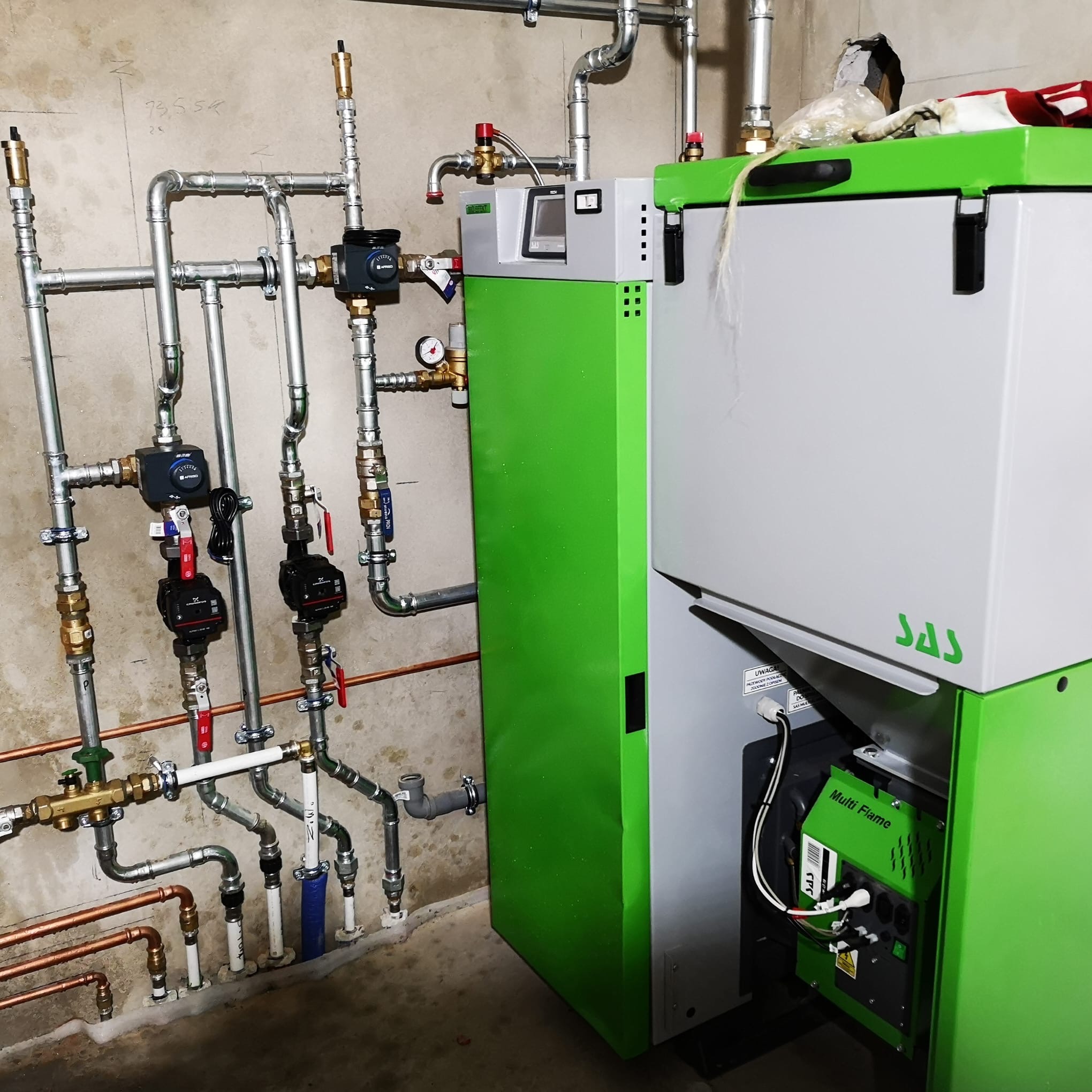 Nowoczesny, zielono-szary piec SAS Multi Flame z podłączoną instalacją hydrauliczną z zaworami i pompami obiegowymi na tle surowej ściany.