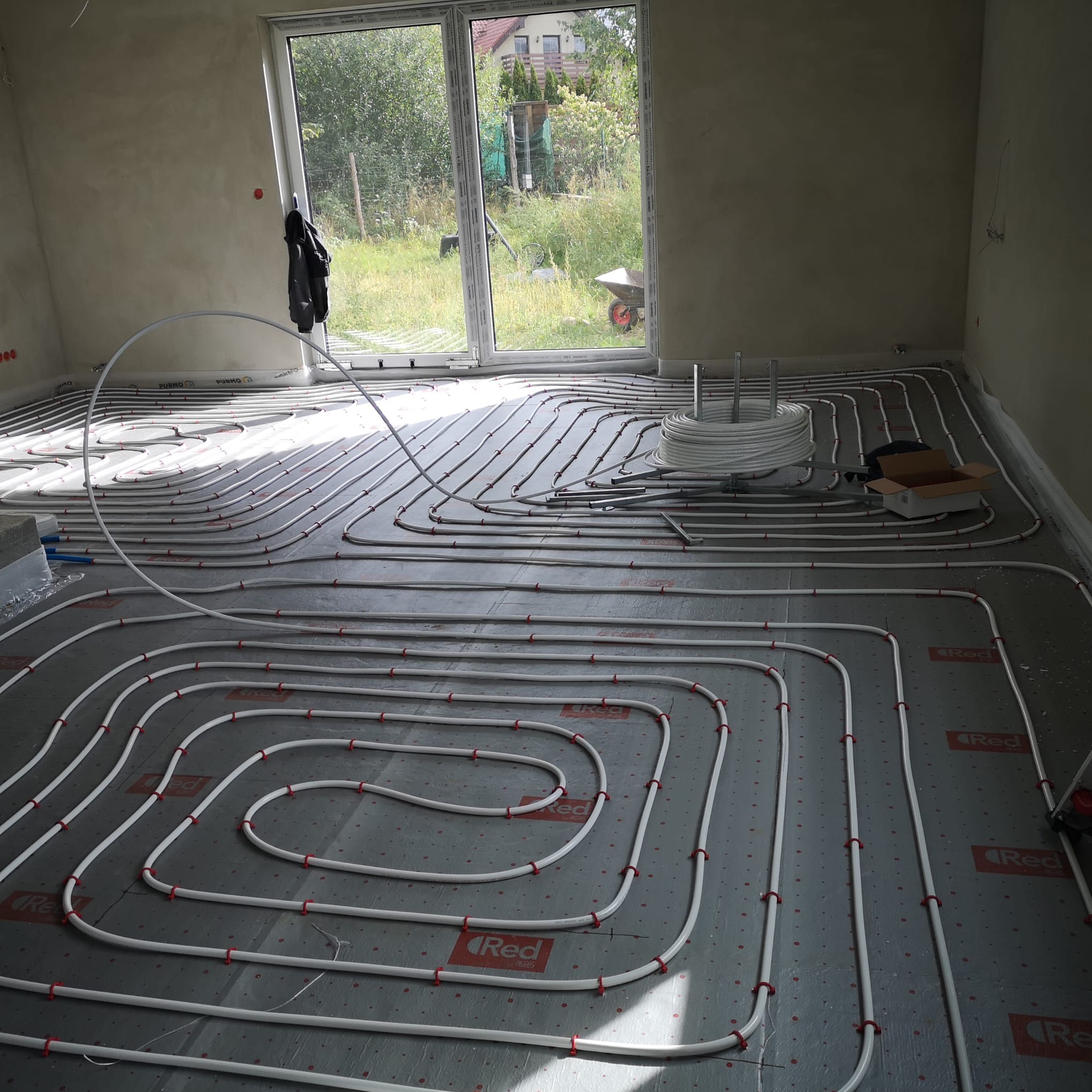 Białe rury ogrzewania podłogowego ułożone na szarej macie z czerwonymi elementami mocującymi, widok z góry na instalację w surowym wnętrzu z oknem.