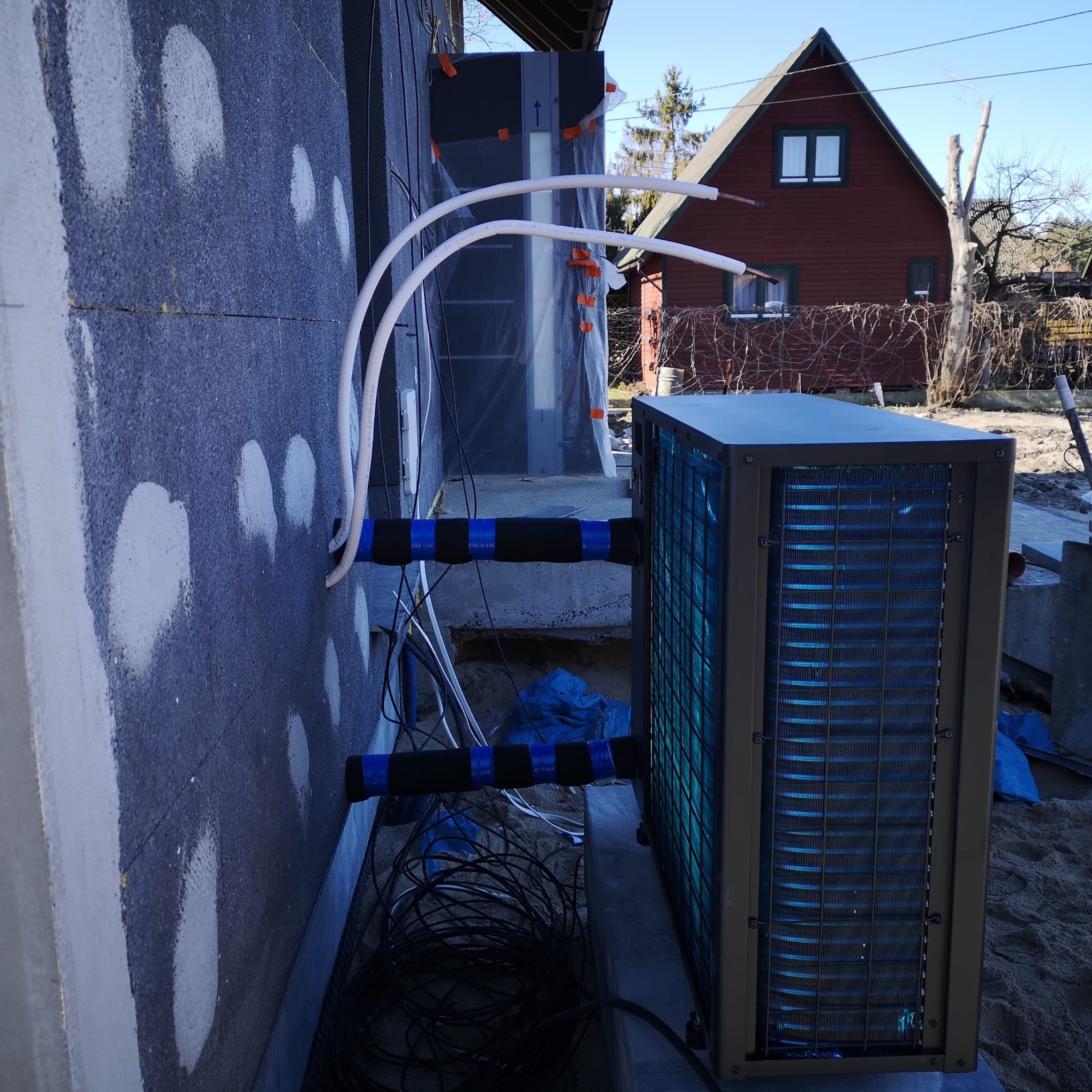 Zewnętrzna jednostka klimatyzacji zamontowana na ścianie budynku z widocznymi rurami i izolacją. W tle dom jednorodzinny i błękitne niebo.