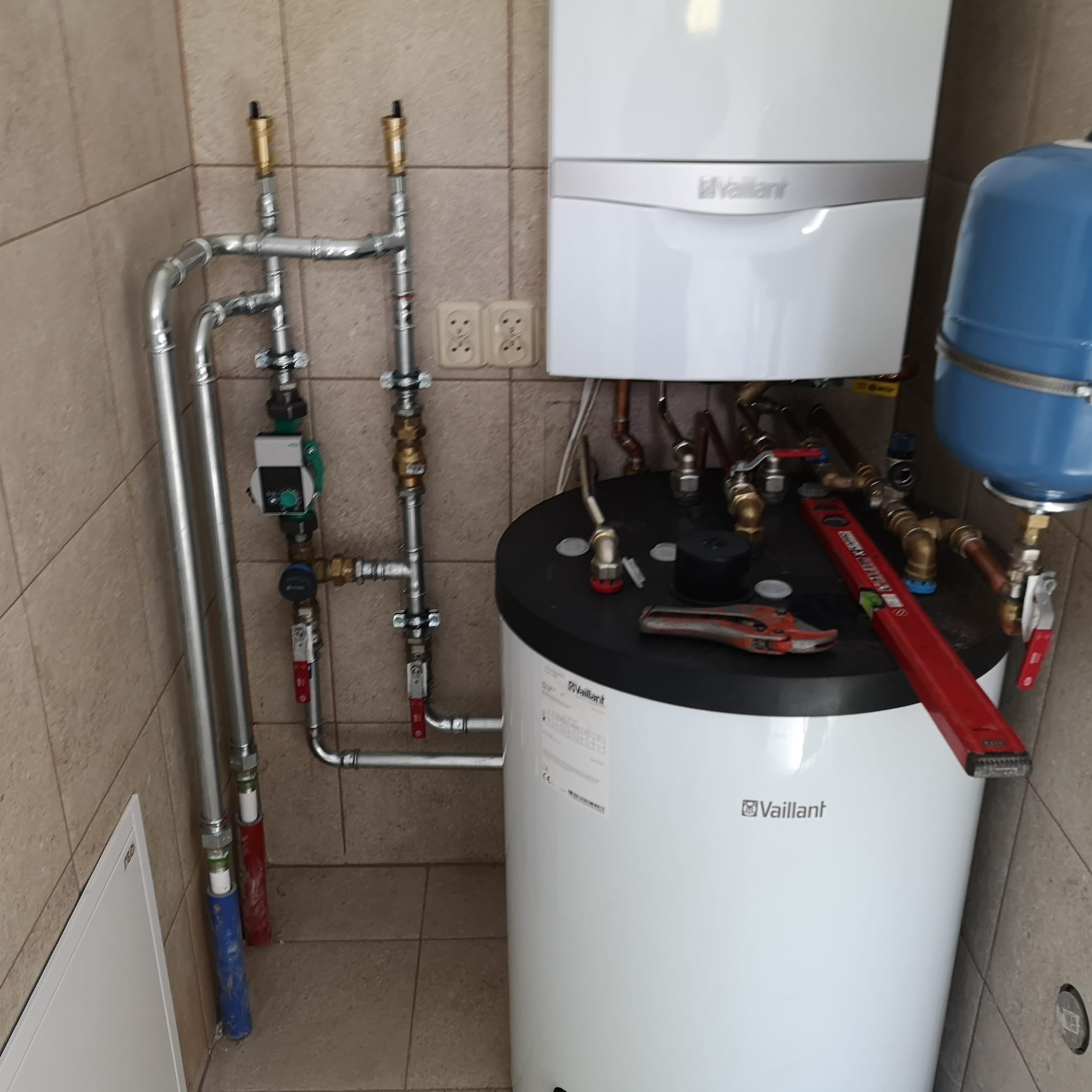 Biały piec gazowy Vaillant z niebieskim zbiornikiem wyrównawczym, połączony rurami. Widoczny poziomica i klucz do rur. Beżowe płytki na ścianach.
