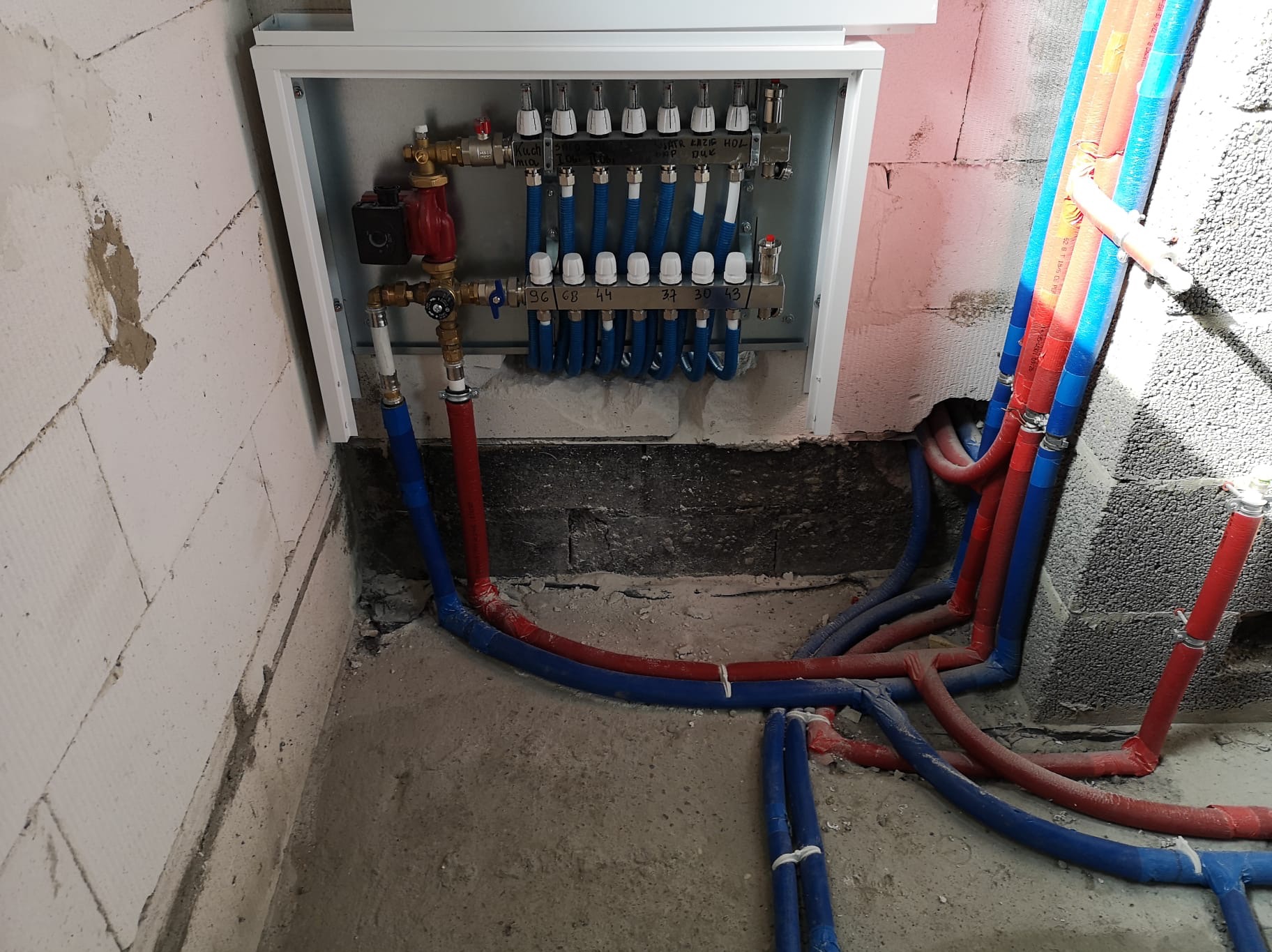 Rozdzielacz ogrzewania podłogowego w białej skrzynce, z podłączonymi rurami w kolorze niebieskim i czerwonym, zamocowanymi do ściany z betonu komórkowego.