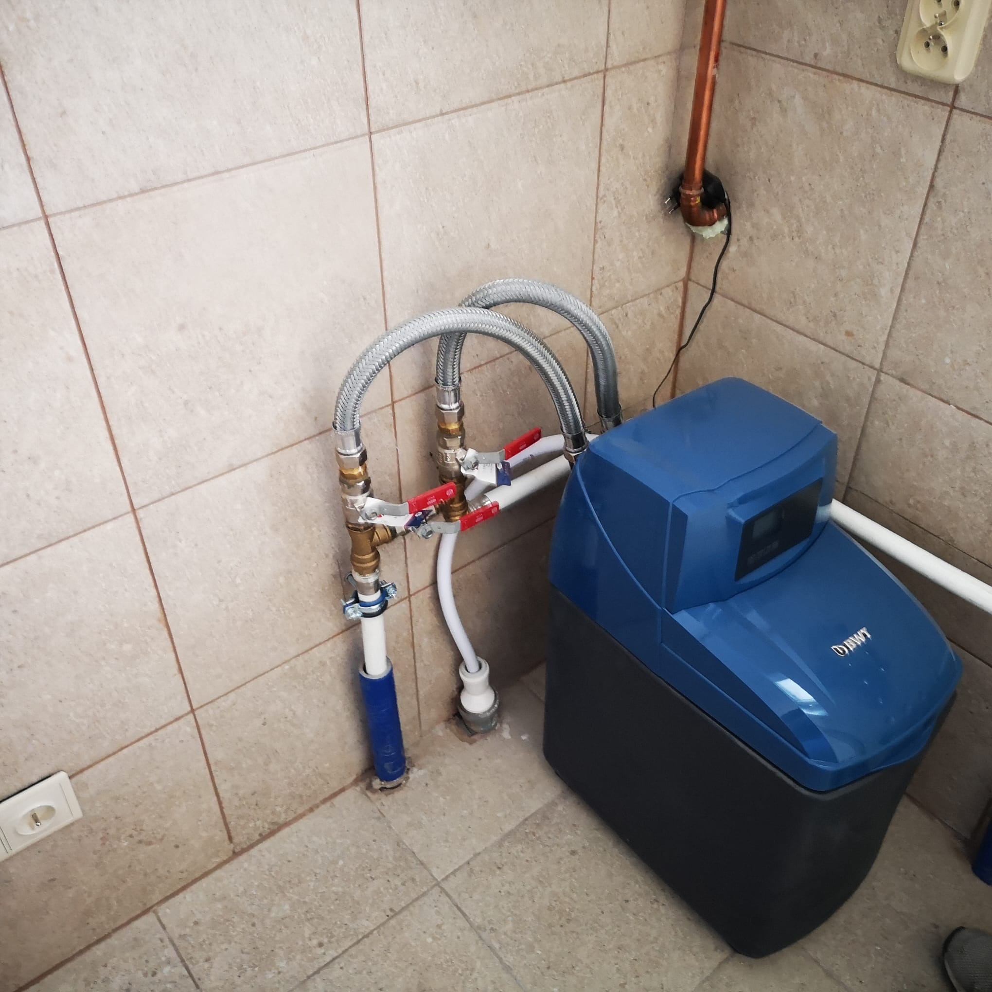 Niebieski zmiękczacz wody BWT podłączony do instalacji hydraulicznej w pomieszczeniu z beżowymi płytkami. Widoczne zawory, węże i rury miedziane.