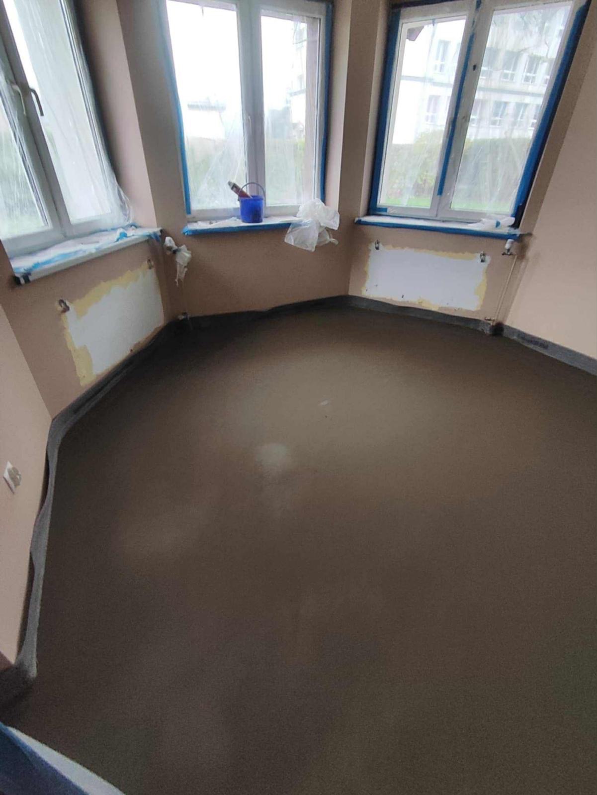 Świeżo wylana, gładka posadzka jastrychowa w pokoju na poddaszu z trzema oknami zabezpieczonymi folią malarską. Widoczne ślady po grzejnikach.