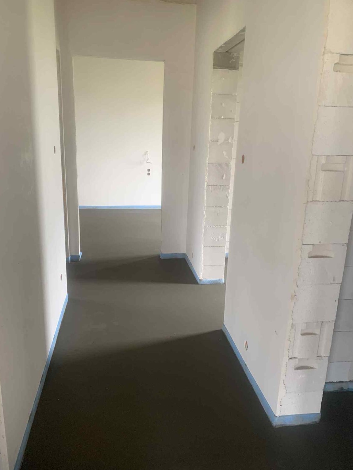 Wnętrze w trakcie prac: świeżo wylana, ciemna posadzka z jastrychu, zabezpieczona niebieską taśmą przy ścianach z białych bloczków. Widok z perspektywy korytarza.