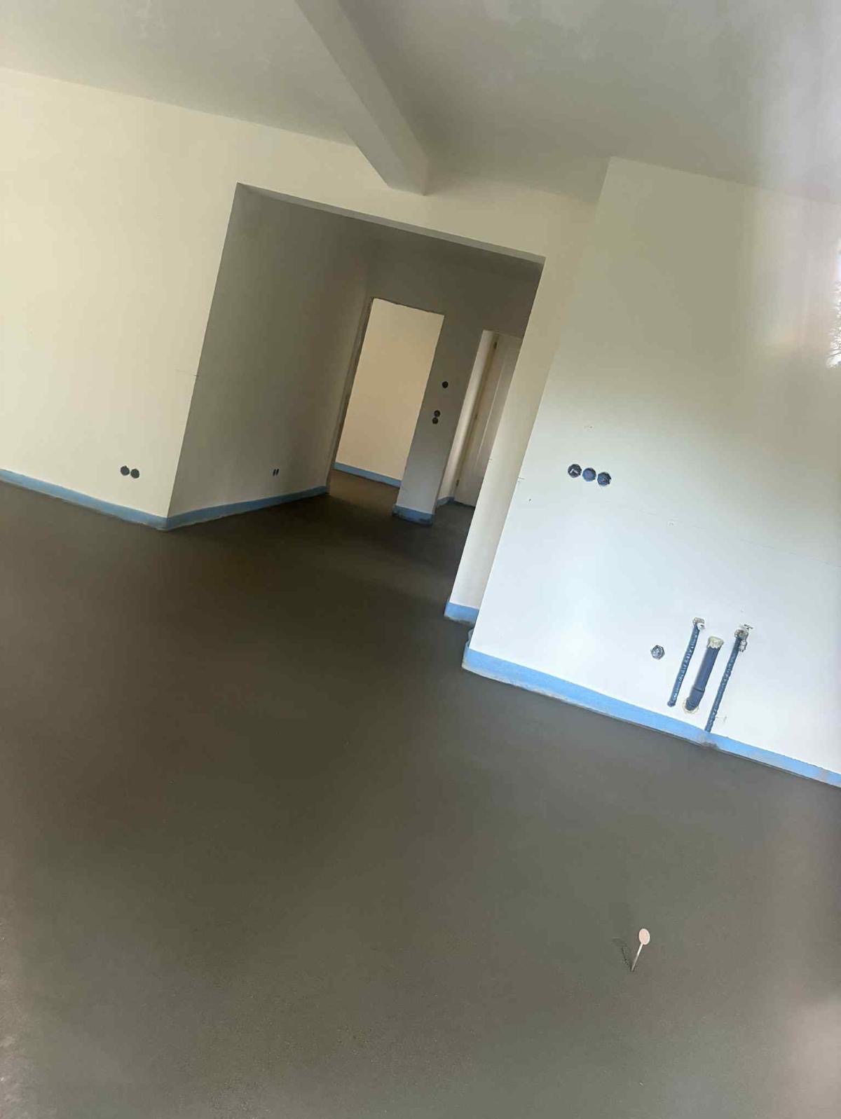 Świeżo wylana, gładka posadzka jastrychowa w jasnym pomieszczeniu z zabezpieczonymi ścianami. Widoczne wystające rury instalacyjne i otwory elektryczne.