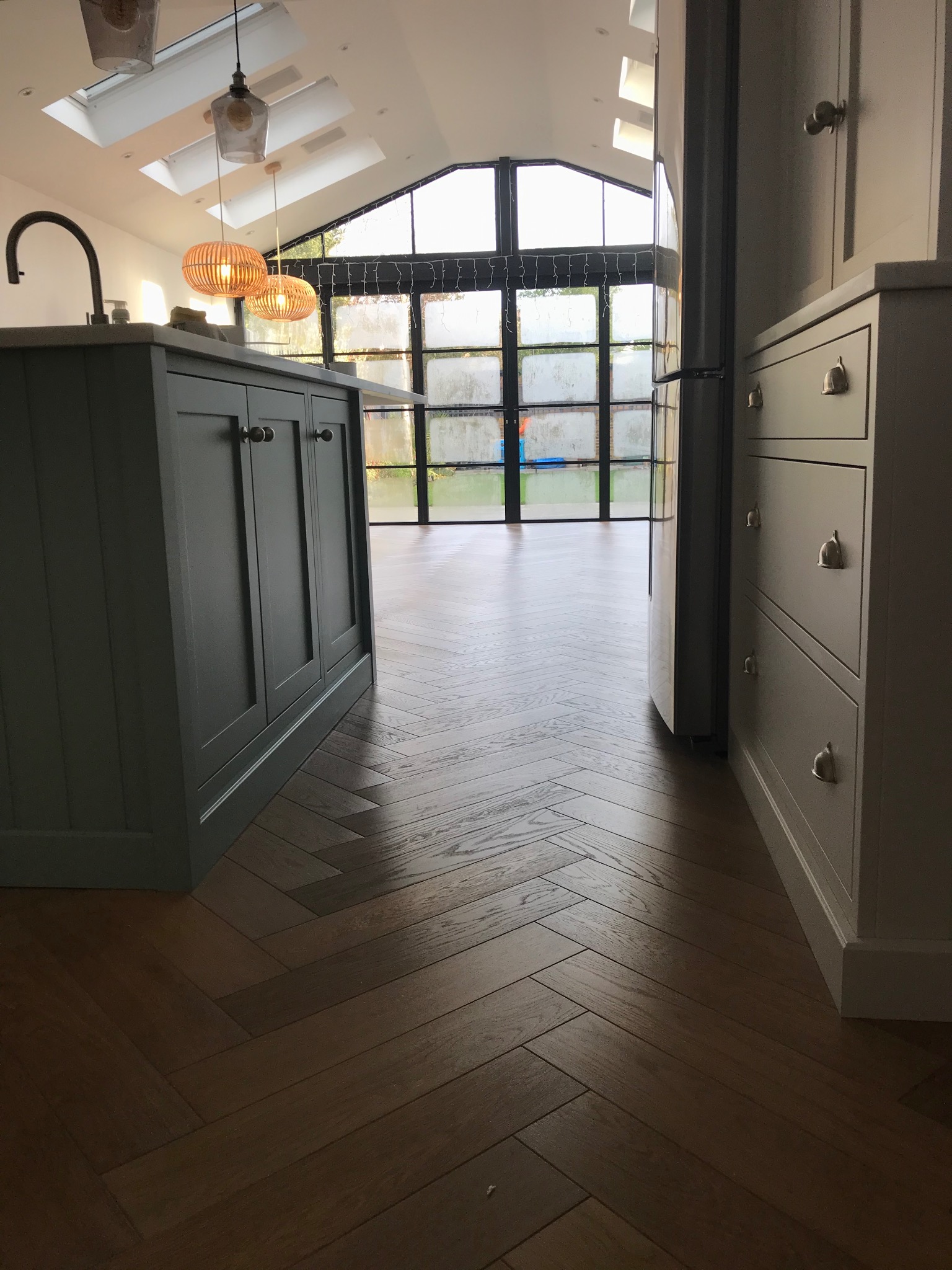 Ukończona podłoga z parkietu ułożonego w jodełkę w nowoczesnej kuchni z wyspą i dużym oknem. Widoczne detale wykończenia i oświetlenie.