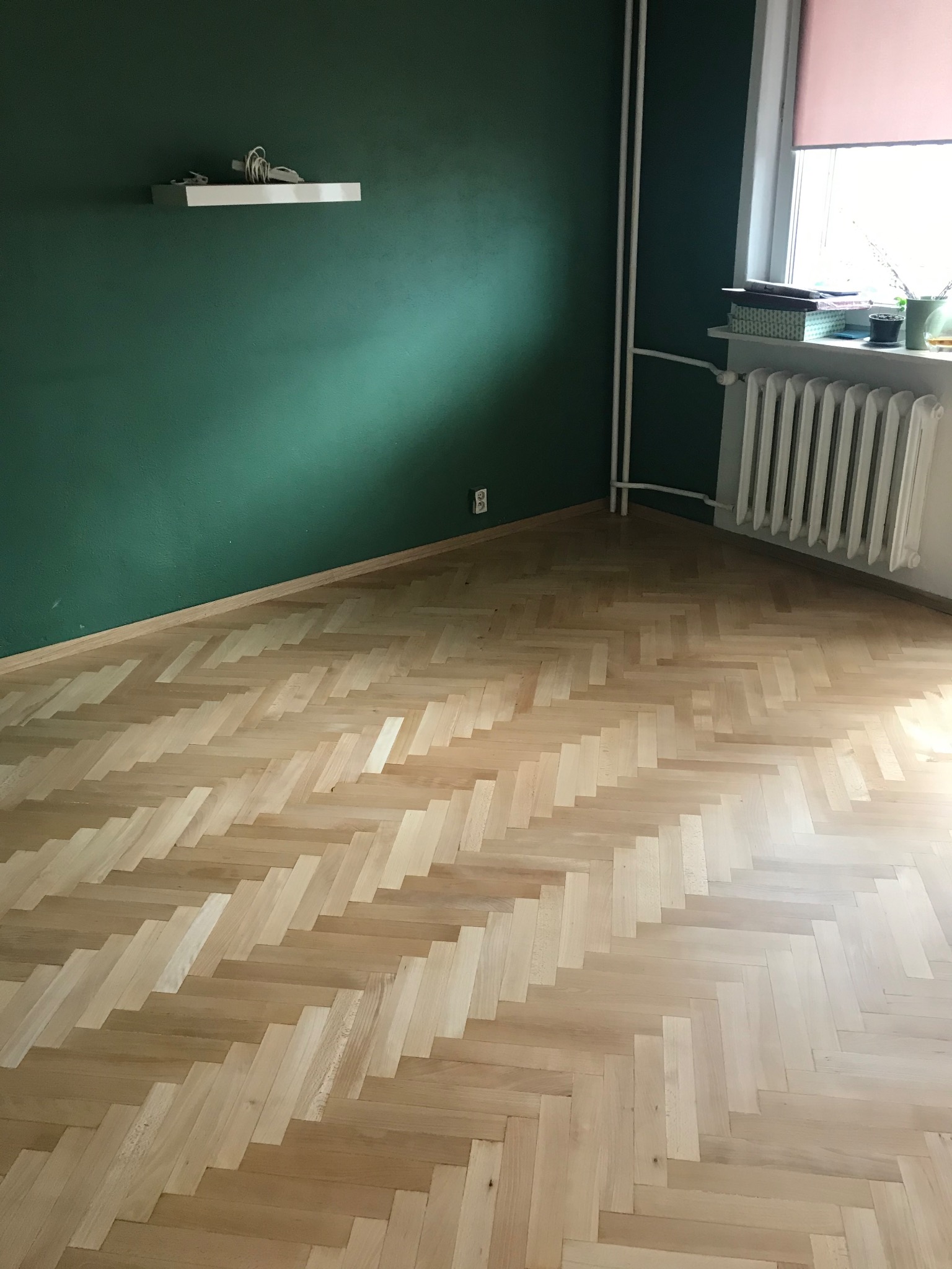 Jasny parkiet ułożony w jodełkę na tle ściany w odcieniach zieleni. Widoczne białe rury grzejne i fragment okna z roletą. Minimalistyczna półka na ścianie.