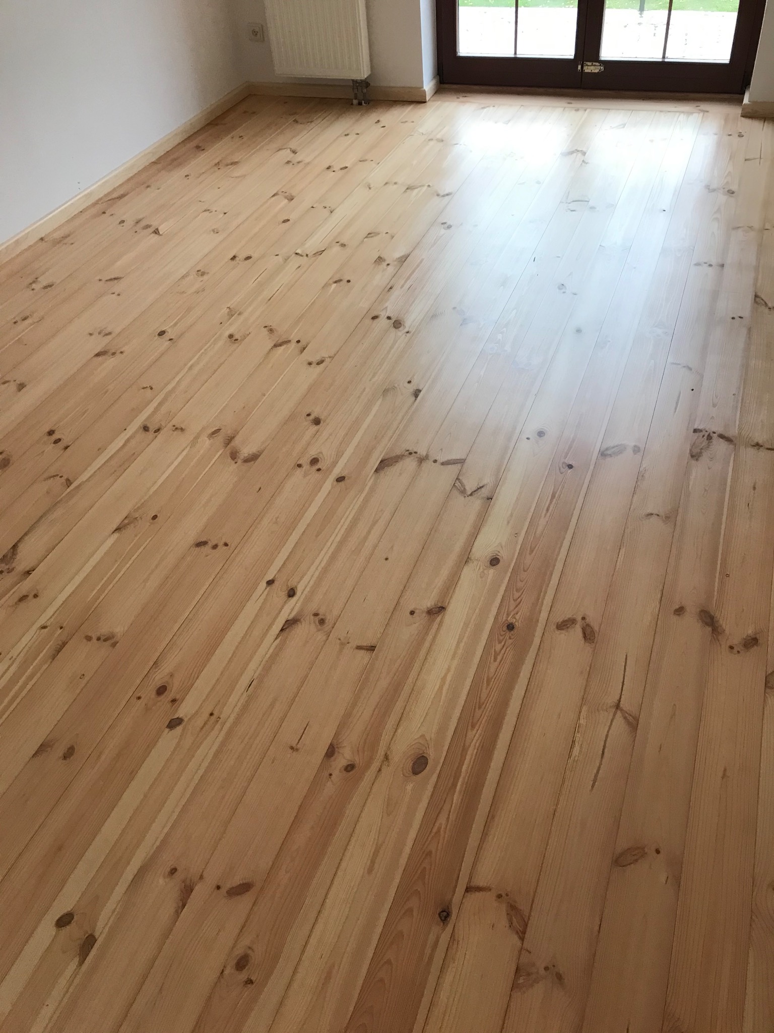 Jasne, drewniane deski podłogowe z widocznymi słojami, ułożone równolegle w przestronnym pokoju z białymi ścianami i oknem balkonowym w tle.