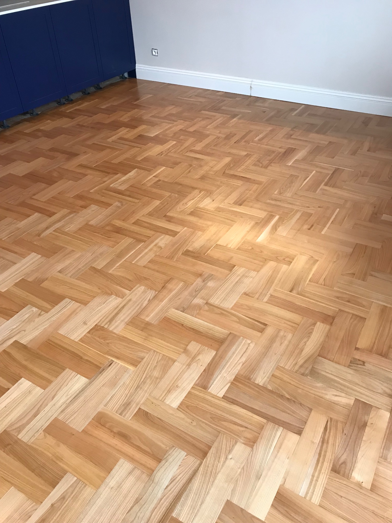 Ułożony parkiet w jodełkę z jasnego drewna, widok z góry, fragment niebieskiej szafki w tle, biała listwa przypodłogowa i gniazdko elektryczne na ścianie.