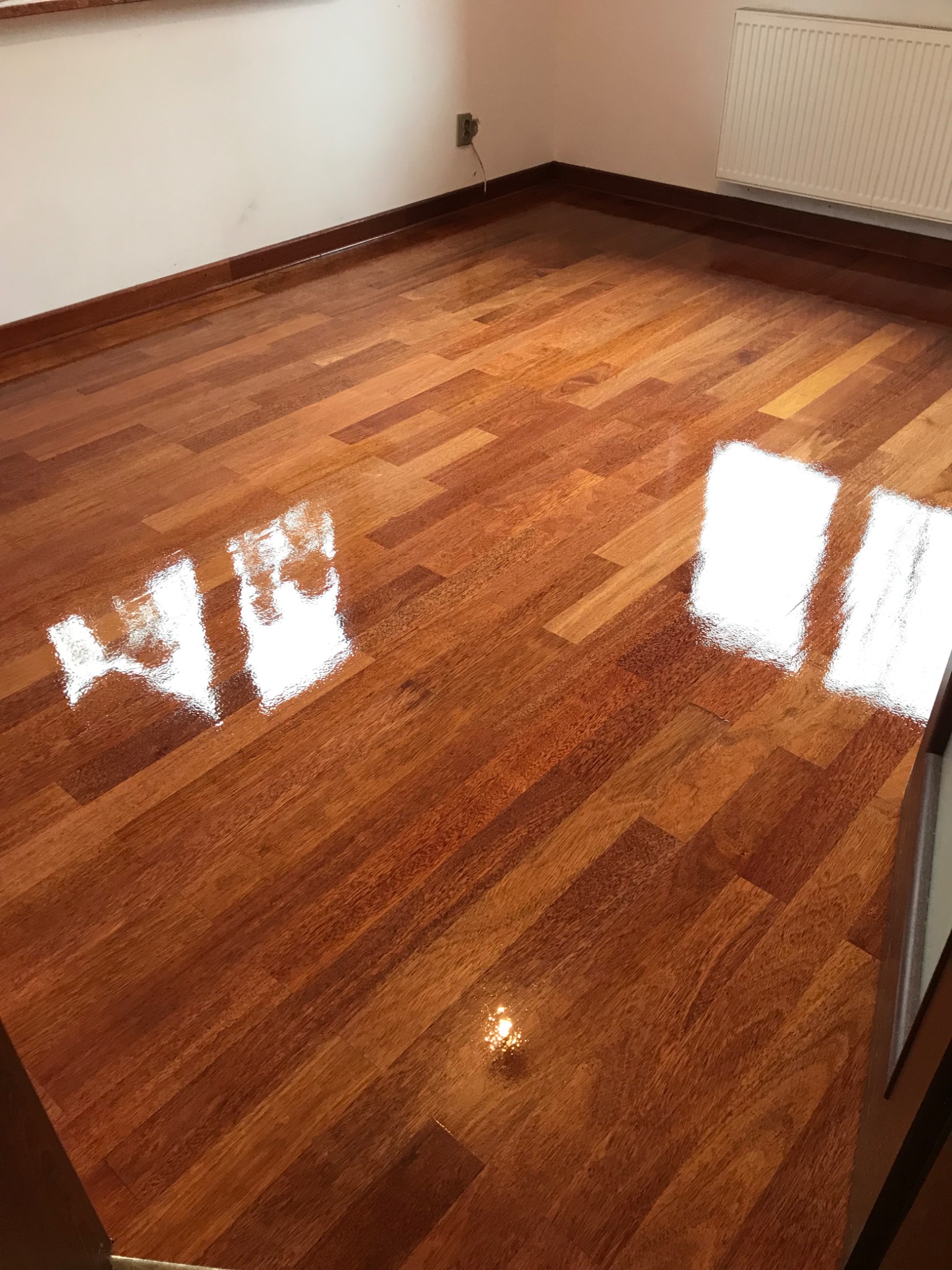 Błyszcząca, świeżo polakierowana podłoga parkietowa z egzotycznego drewna w pokoju, odbijająca światło okien. Widoczne listwy przypodłogowe i fragment grzejnika.