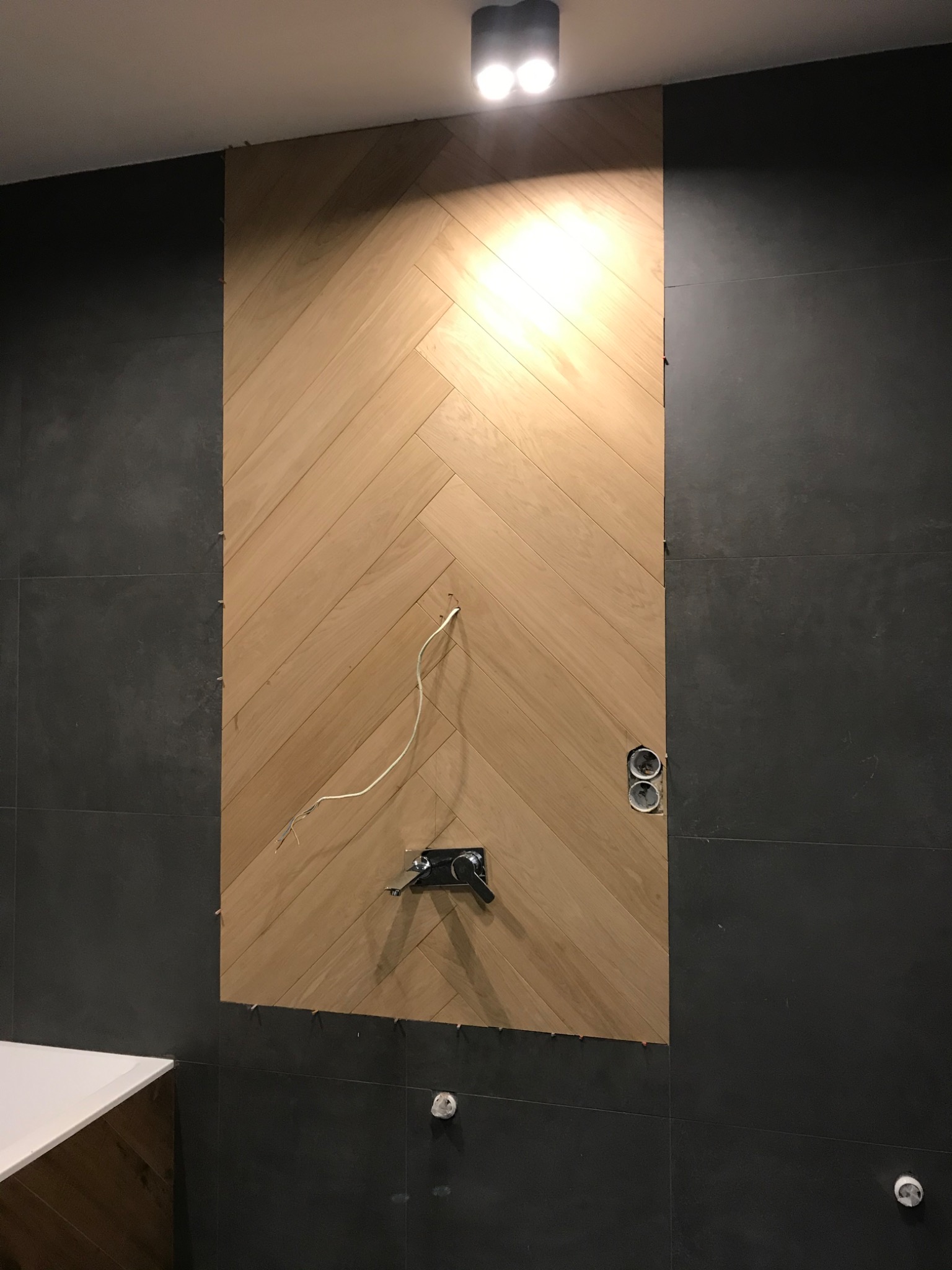 Ściana z parkietu ułożonego w jodełkę, z widocznymi elementami instalacji wodnej i elektrycznej. Ciemne płytki po bokach, sufit z oświetleniem.