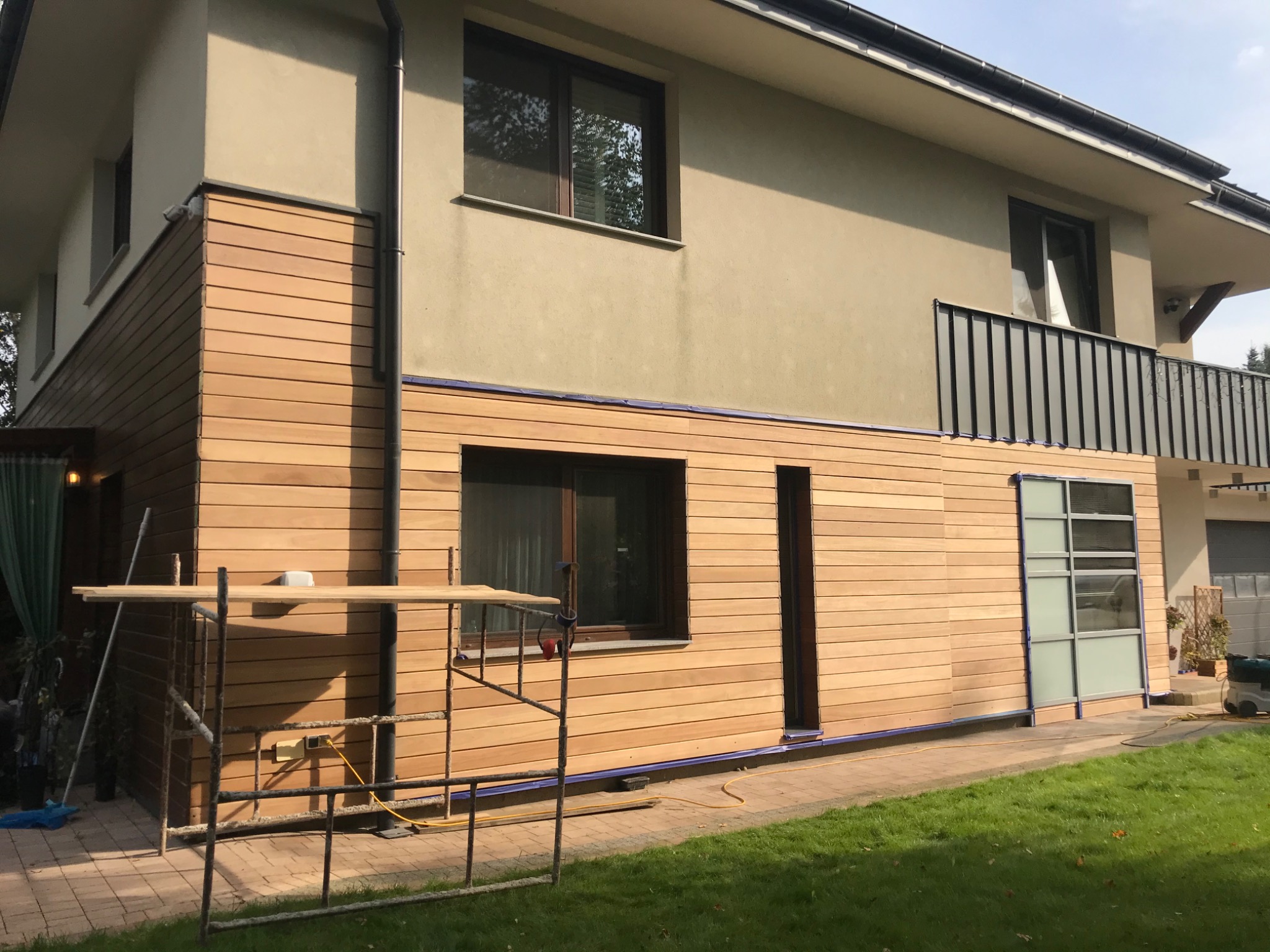 Elewacja domu z drewnianą okładziną, widoczne rusztowanie i narzędzia. Detal: poziomy układ desek, jasne drewno, kontrast z beżową ścianą. Prace wykończeniowe.