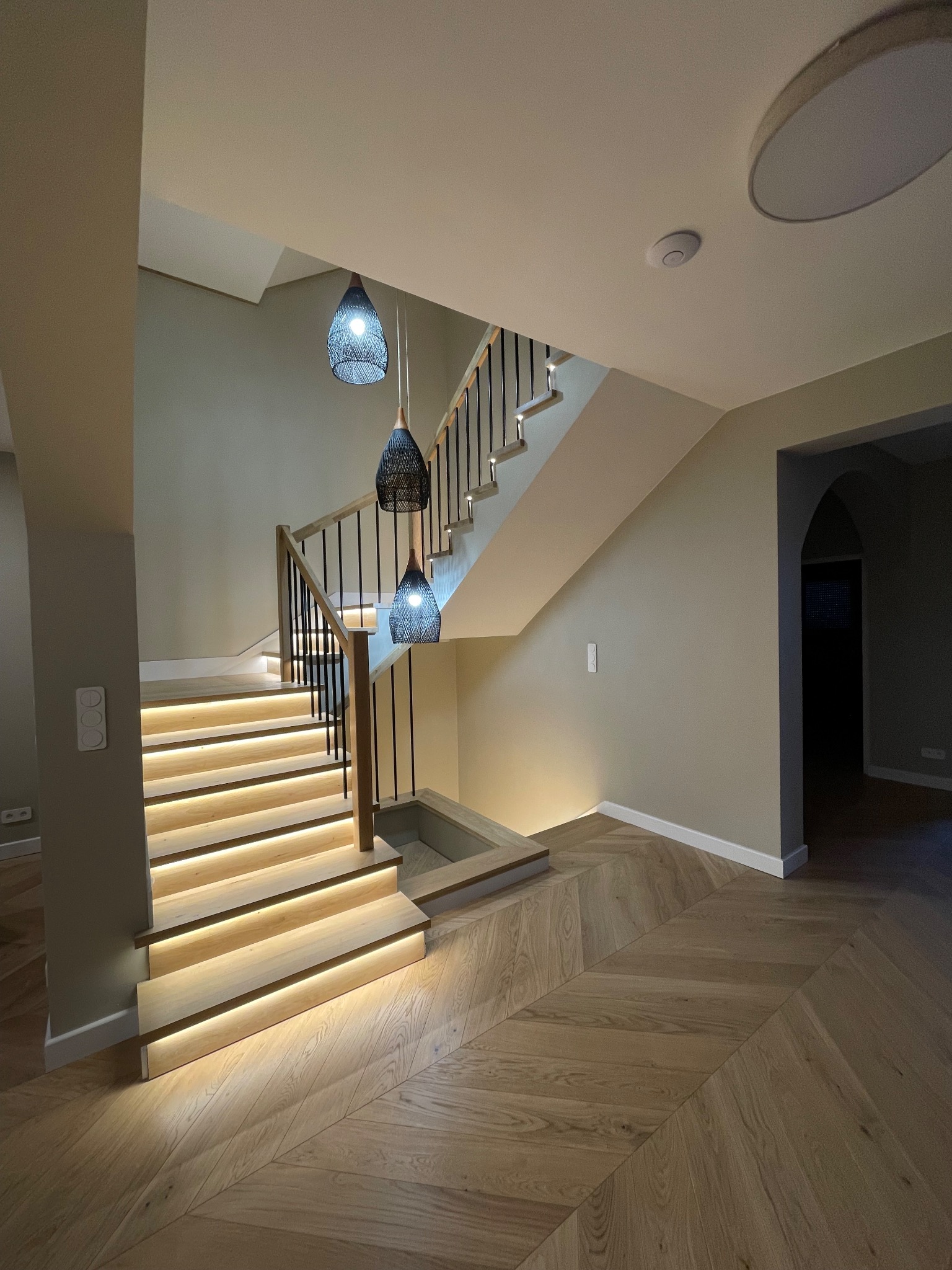 Nowoczesne schody z podświetleniem LED i parkietem ułożonym we wzór jodełki. Czarne balustrady i drewniane stopnie tworzą elegancki design wnętrza.