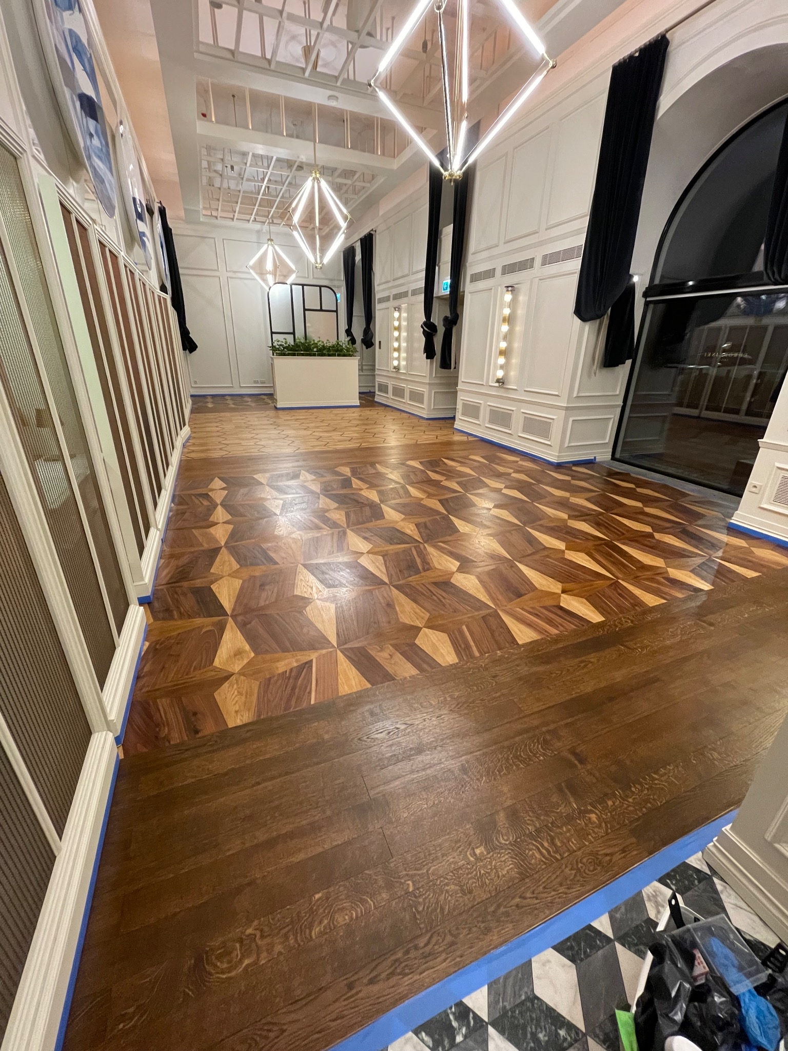 Eleganckie wnętrze z parkietem w geometryczny wzór, zabezpieczonym niebieską taśmą malarską. Widoczne oświetlenie i dekoracyjne elementy architektoniczne.