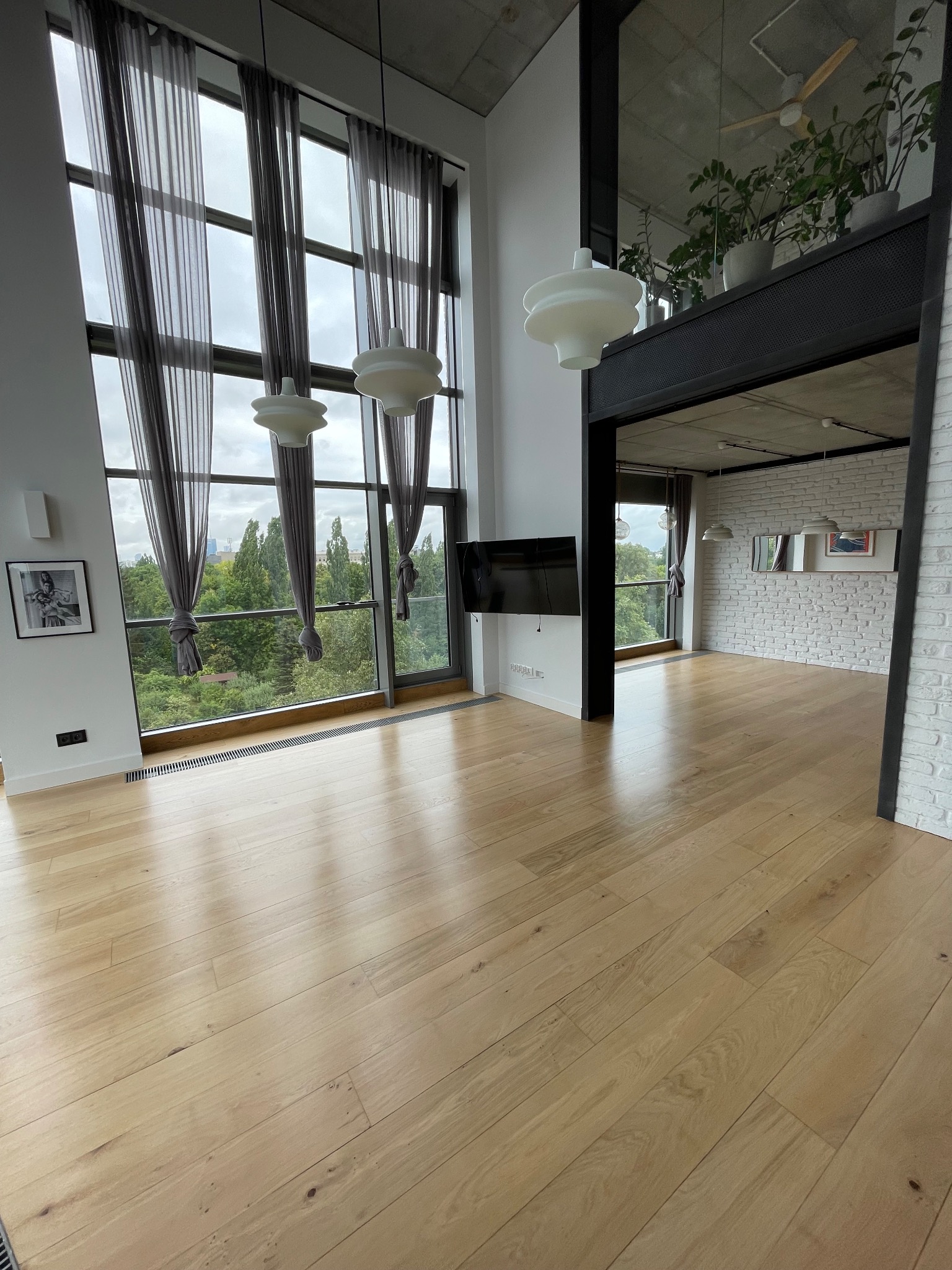 Jasny parkiet w przestronnym, nowoczesnym wnętrzu z dużym oknem i widokiem na zieleń. Minimalistyczne lampy wiszące i biała cegła dodają elegancji.