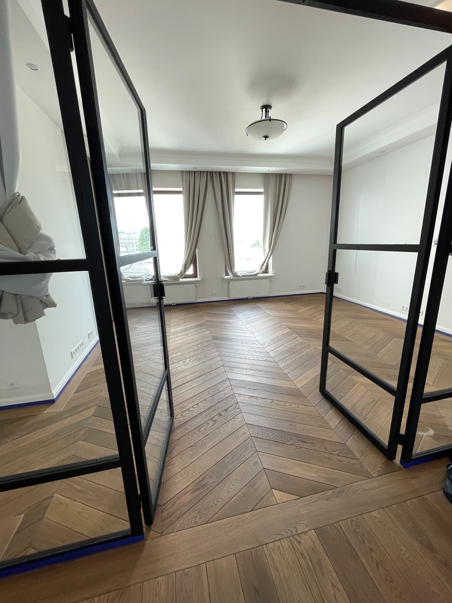 Parkiet ułożony w jodełkę francuską, widoczny przez otwarte, czarne, przeszklone drzwi. Podłoga zabezpieczona niebieską taśmą malarską przy ścianach.