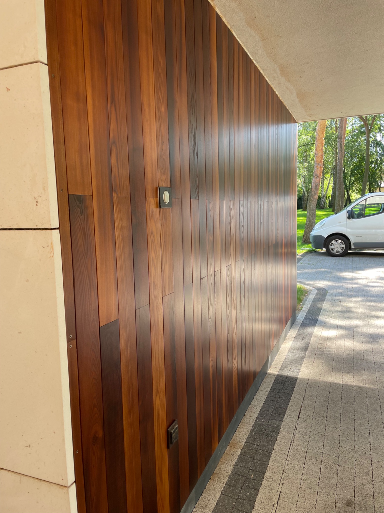 Elegancka, pionowa boazeria z ciemnego drewna na elewacji budynku, z widocznymi detalami montażu i elementami instalacji elektrycznej. W tle fragment chodnika i zieleń.