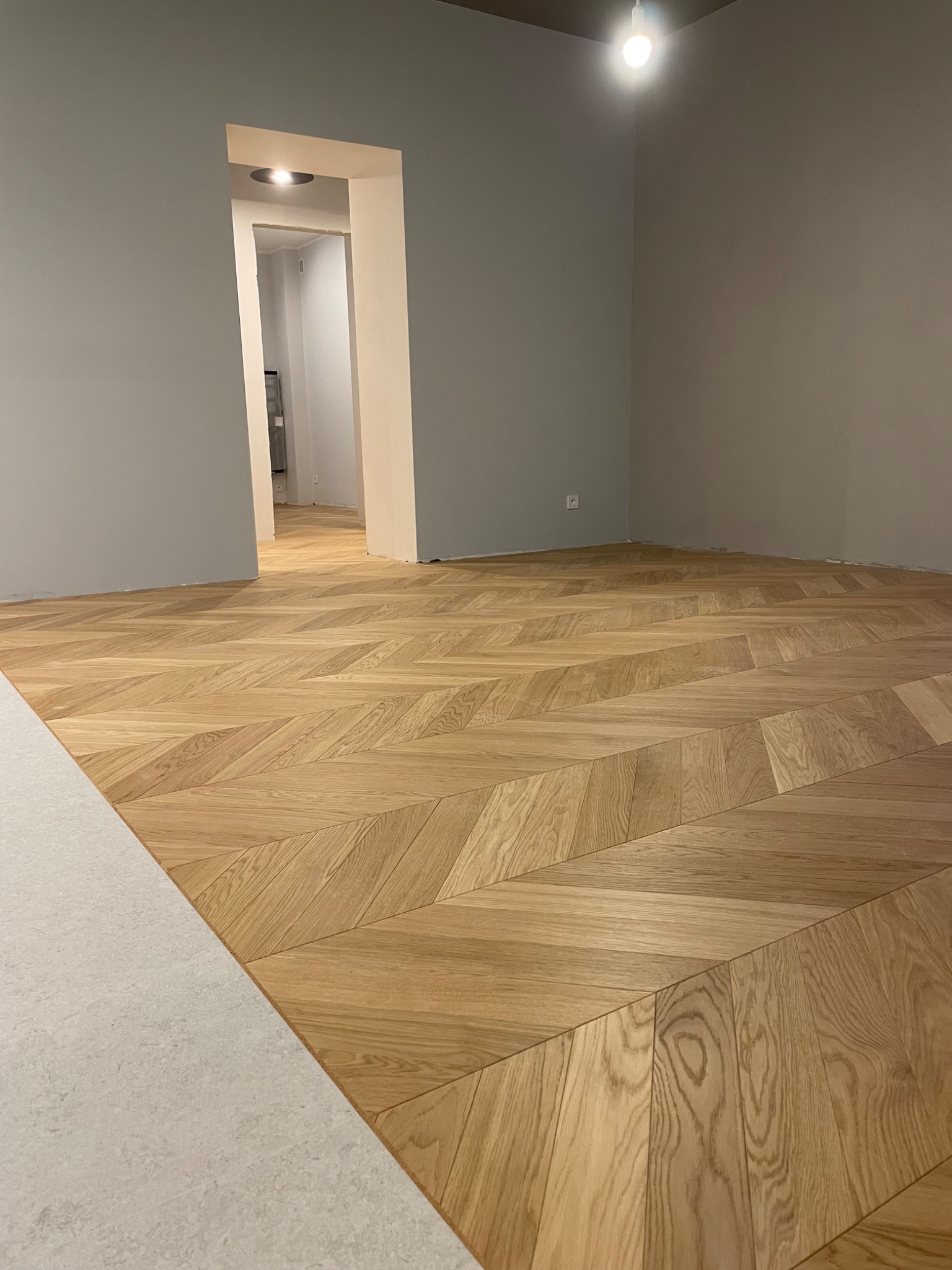 Jasny parkiet ułożony we wzór jodełki w nowym, pustym pokoju z szarymi ścianami i białymi futrynami drzwi. Widok na sąsiednie pomieszczenie.