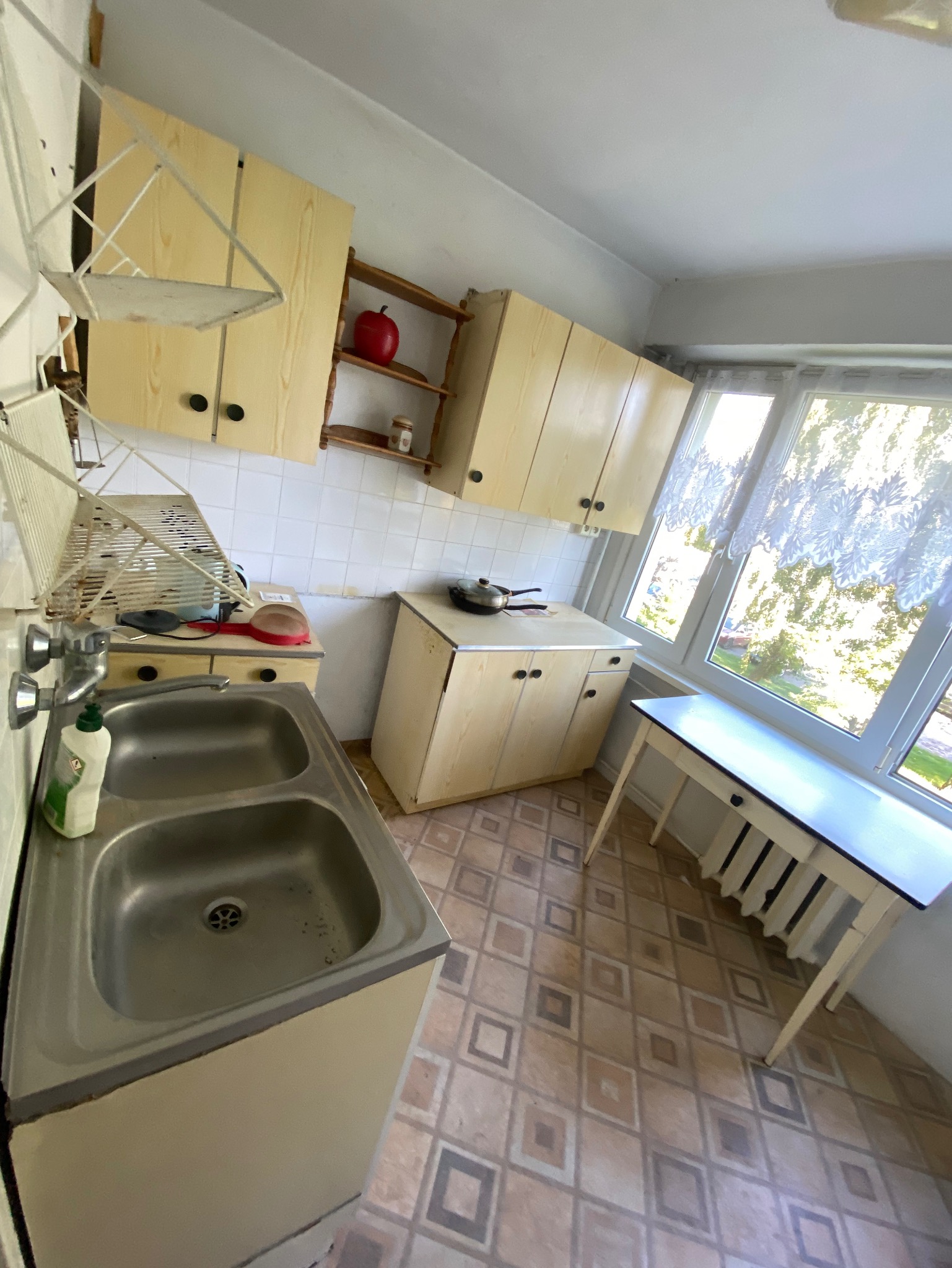 Kuchnia z lat 80. z beżowymi szafkami, metalowym zlewozmywakiem i stołem przy oknie. Na półce czerwone jabłko. Podłoga w kwadraty. Widok z góry.