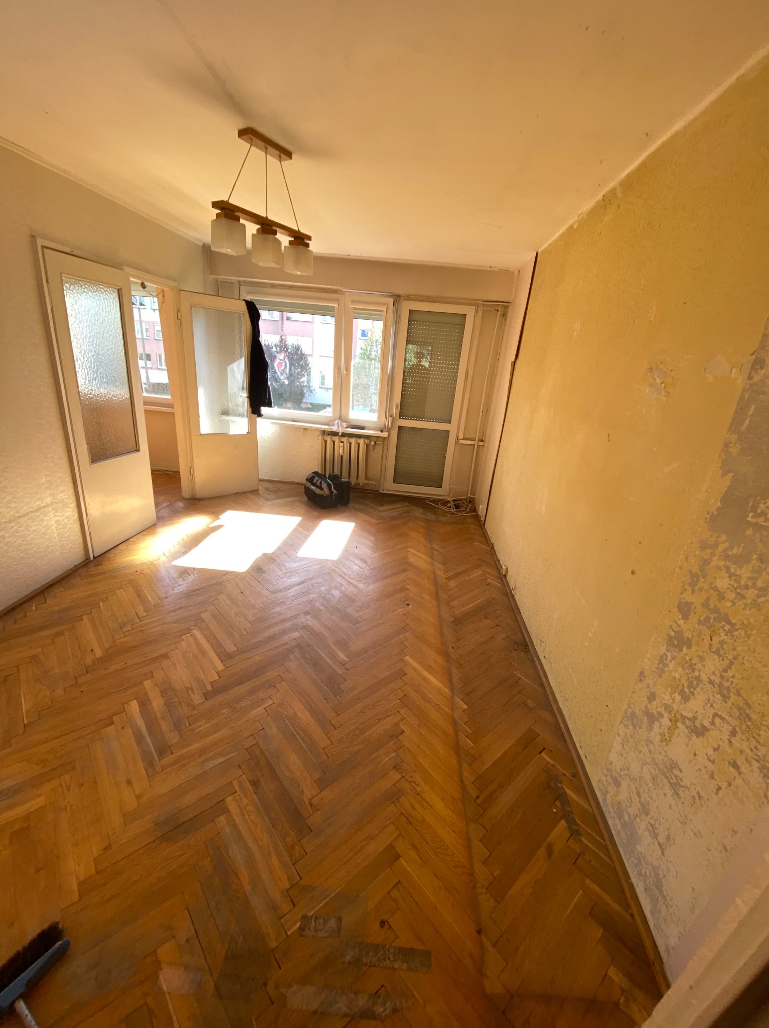 Pokój po remoncie: parkiet w jodełkę, ściany z resztkami farby, otwarte drzwi balkonowe, widoczny grzejnik i torba. Przygotowanie do sprzątania.