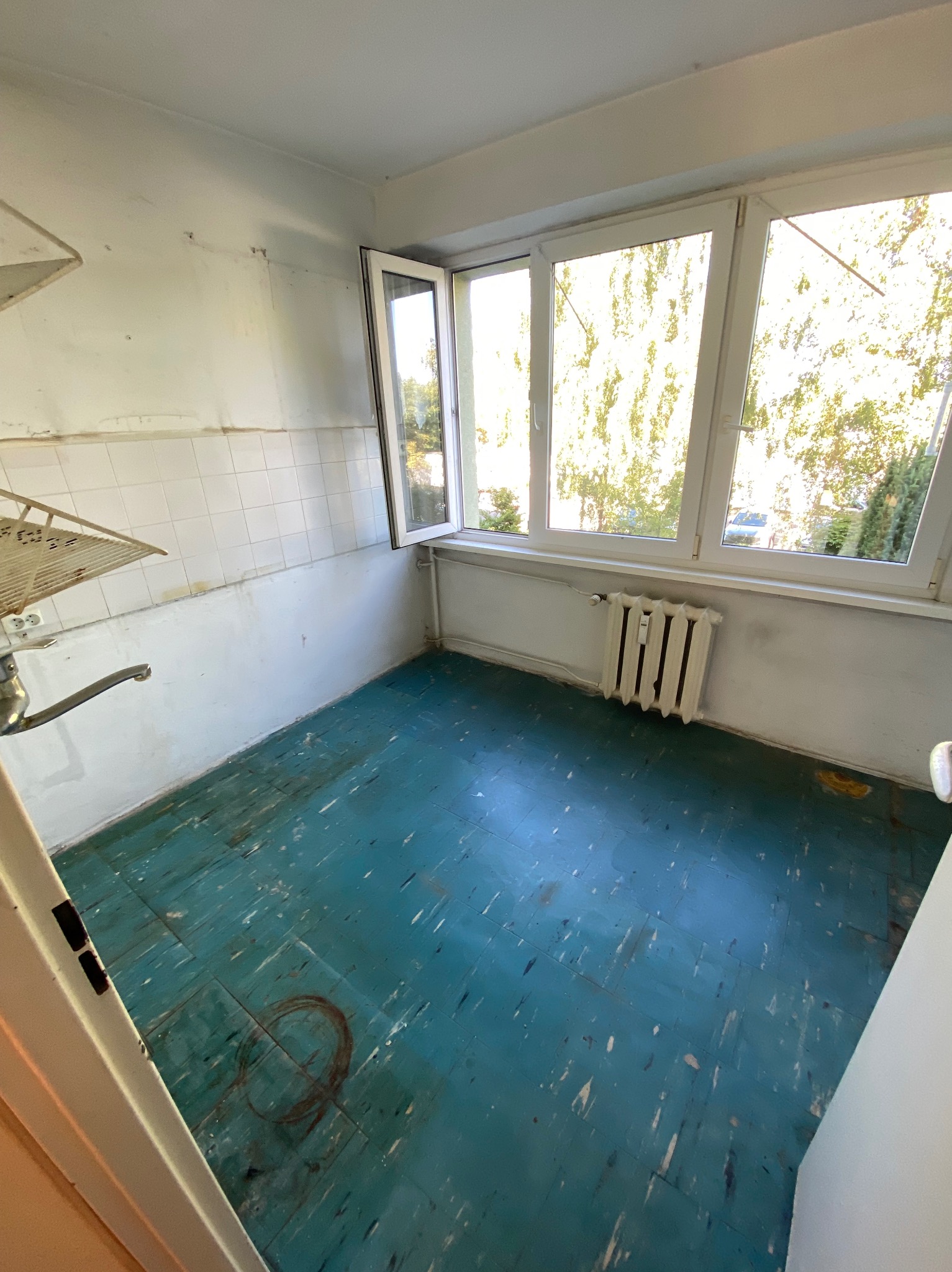 Wnętrze zaniedbanego pokoju z niebieską podłogą, białymi ścianami z resztkami płytek i otwartym oknem z widokiem na zieleń. Widoczny stary grzejnik i fragment instalacji.