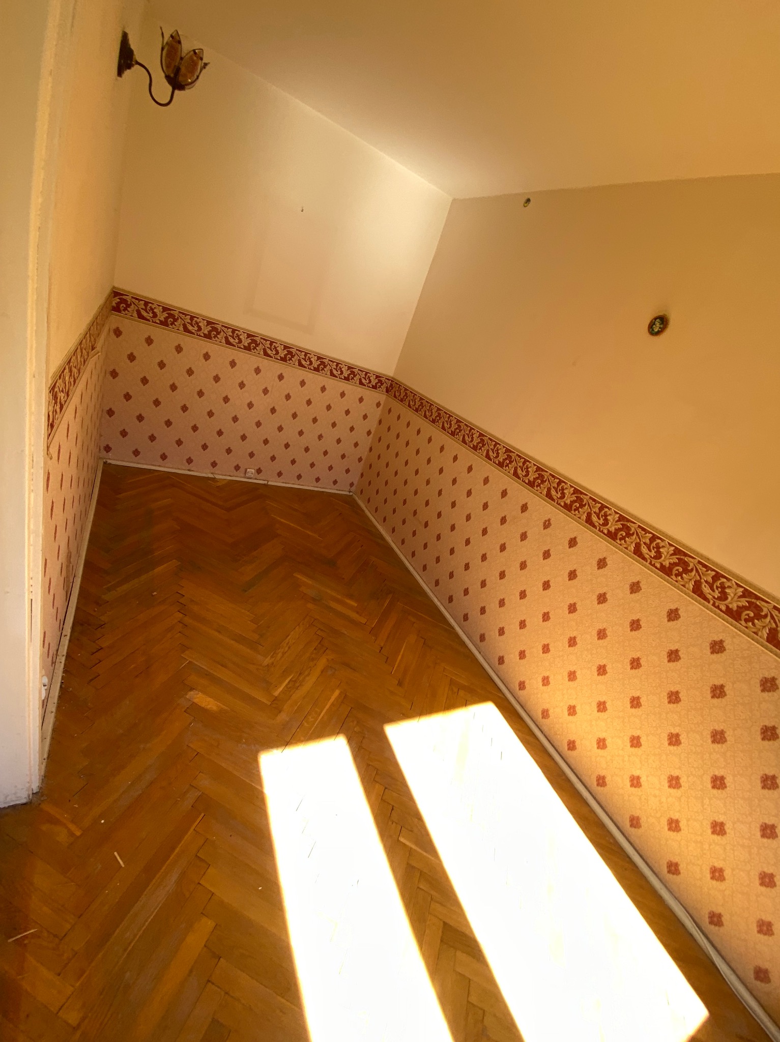Poddasze z parkietem w jodełkę i tapetą w drobne wzory, oświetlone promieniami słońca. Widoczna lampa ścienna i ślady po demontażu na suficie.