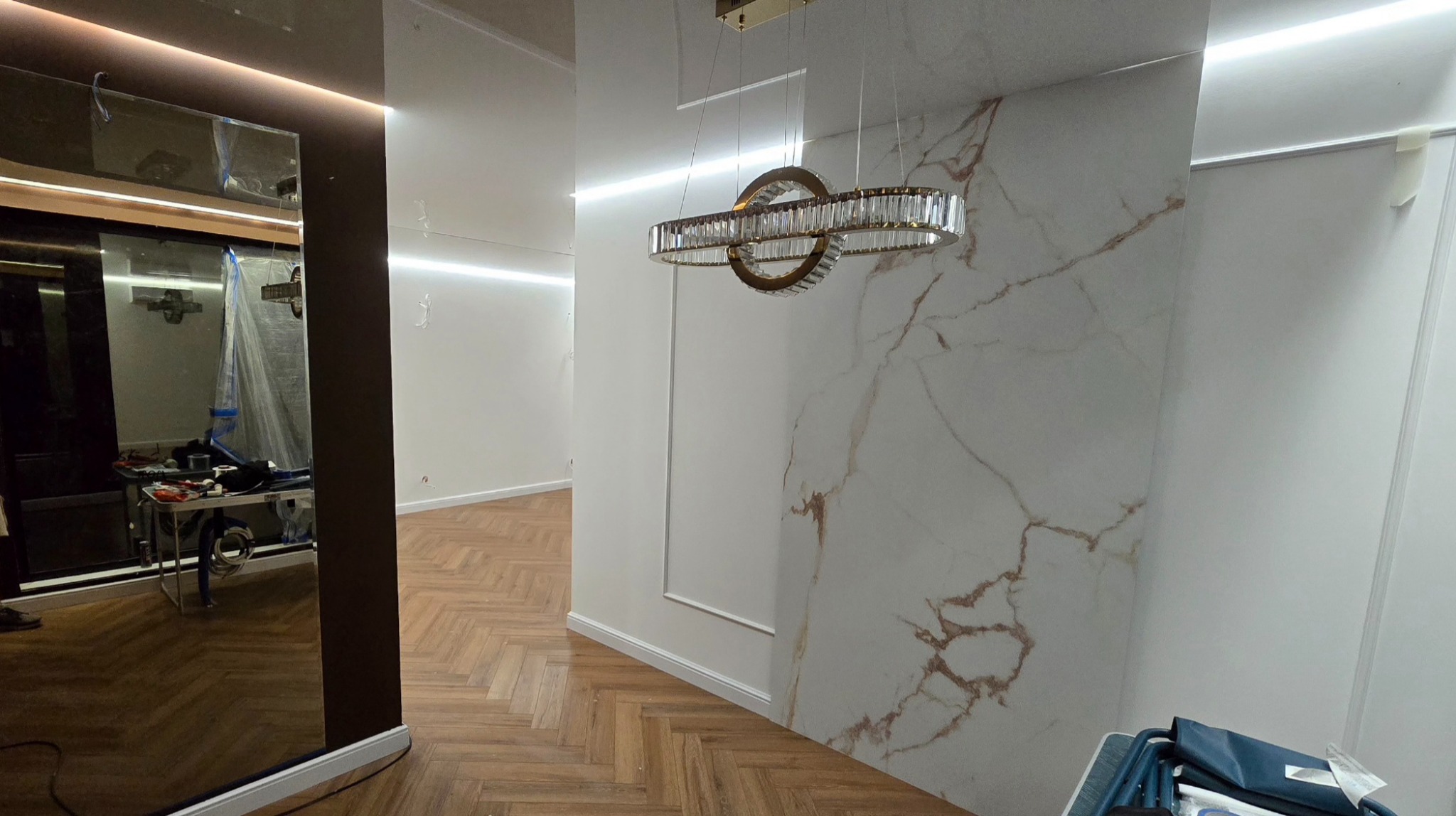 Salon po wykończeniu: parkiet w jodełkę, ściana z marmurowym wzorem, ozdobna lampa. W lustrze odbicie narzędzi i folii ochronnej, sugerujące prace wykończeniowe.