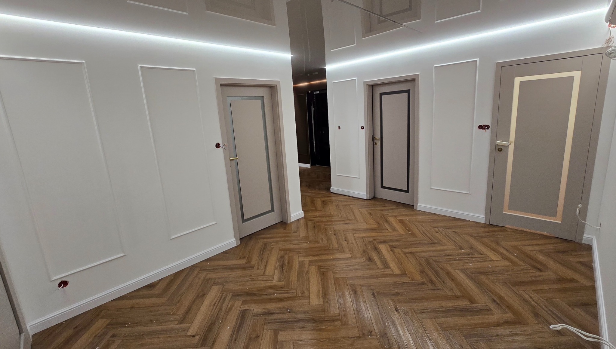 Wykończone wnętrze z parkietem w jodełkę, białymi ścianami z dekoracyjnymi panelami i szarymi drzwiami z ozdobnymi ramkami. Widoczne gniazdka elektryczne.