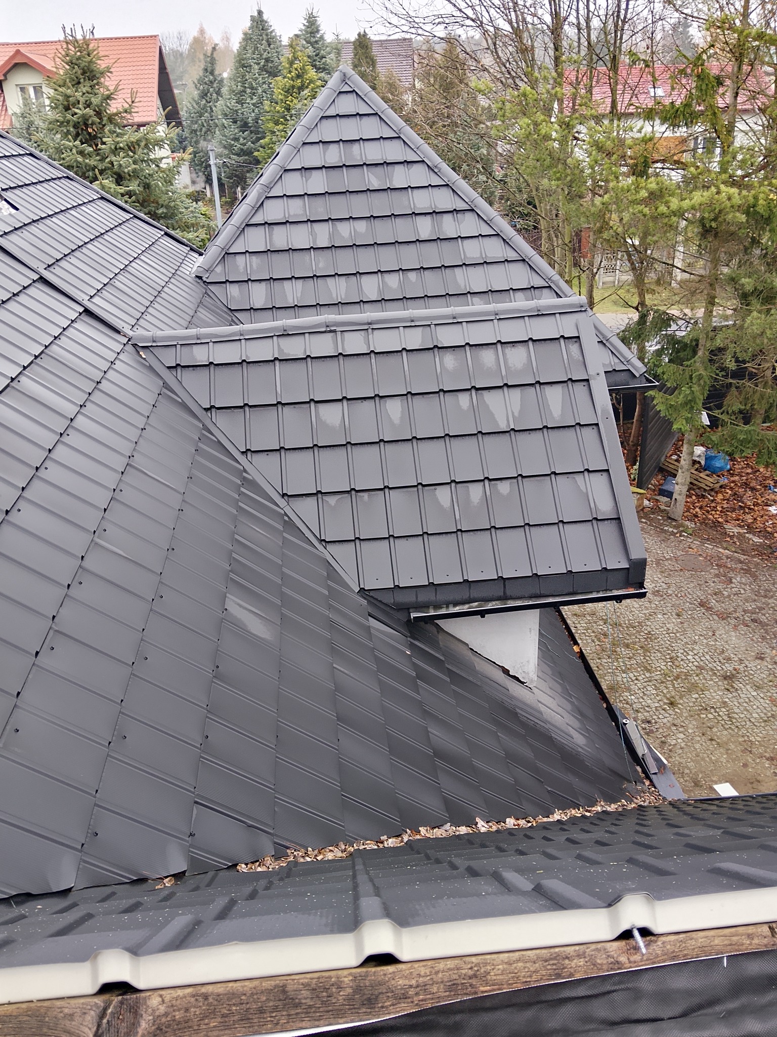 Szary dach z blachy na domu jednorodzinnym, widok z góry, z widocznymi detalami łączeń i fakturą materiału. W tle drzewa i zabudowa mieszkalna.