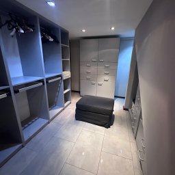 LUXCLEAN LIUBOMYR HRYTSAI - Nowoczesna garderoba z szarymi szafami, półkami i pufą. Minimalistyczny design, jasne płytki podłogowe i punktowe oświetlenie. Widok na uporządkowaną przestrzeń.