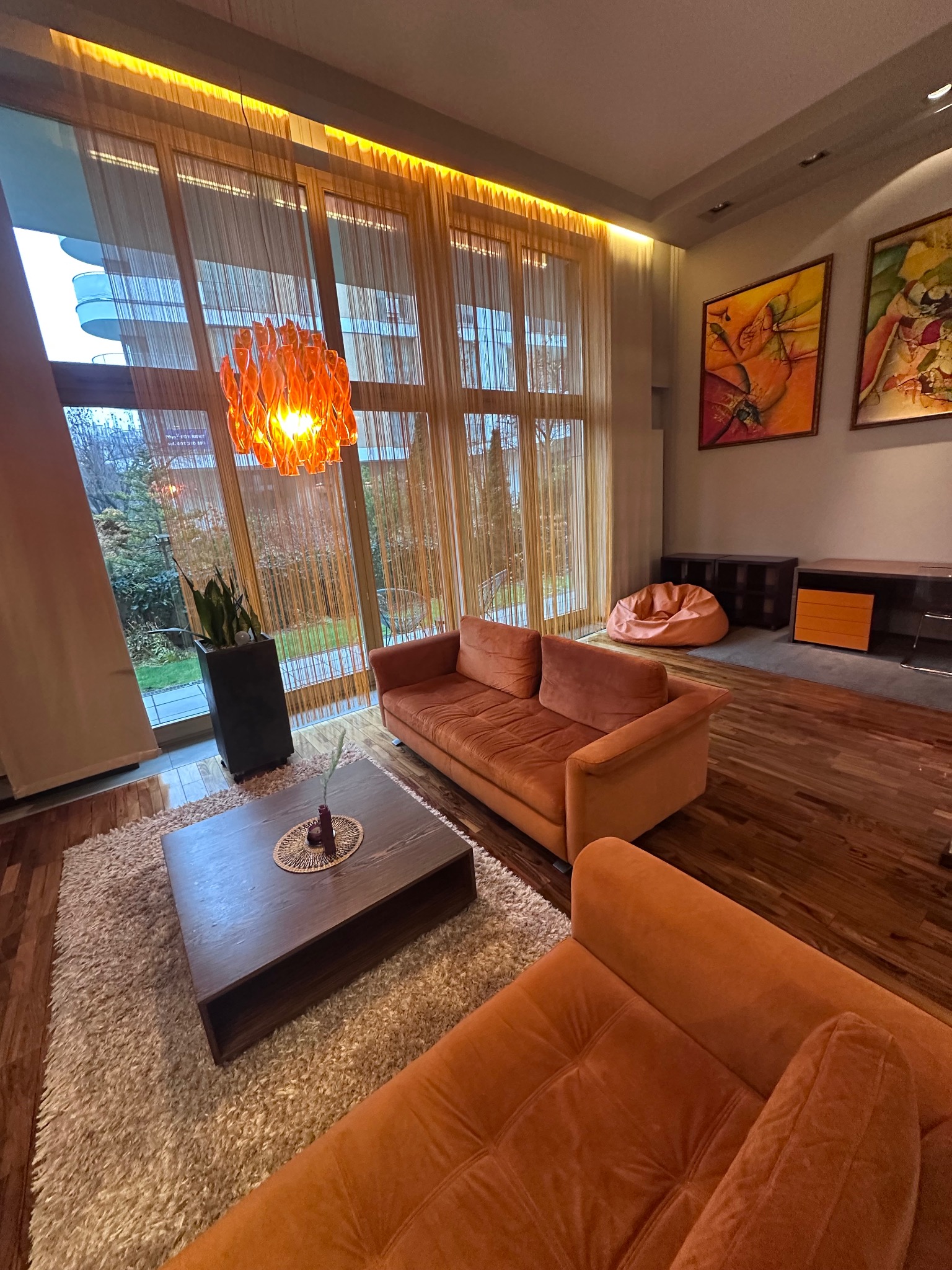 Jasny salon z pomarańczowymi sofami, drewnianą podłogą i dywanem. Duże okno z widokiem na zieleń. Nowoczesny wystrój z obrazami na ścianie i lampą.