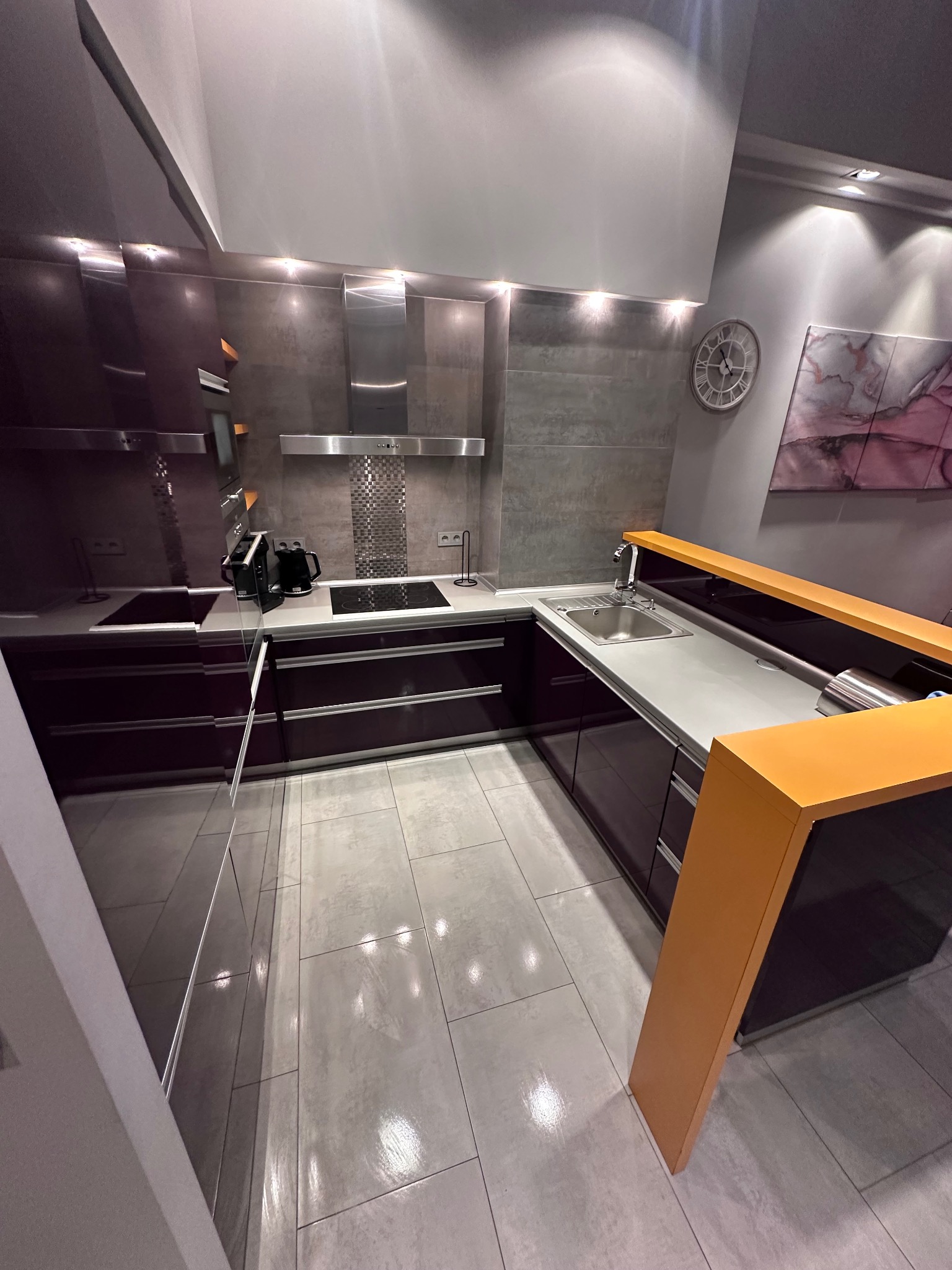 Nowoczesna kuchnia w odcieniach fioletu i szarości z elementami pomarańczowymi. Widok na umywalkę, płytę grzewczą i okap. Na ścianie zegar i obraz.