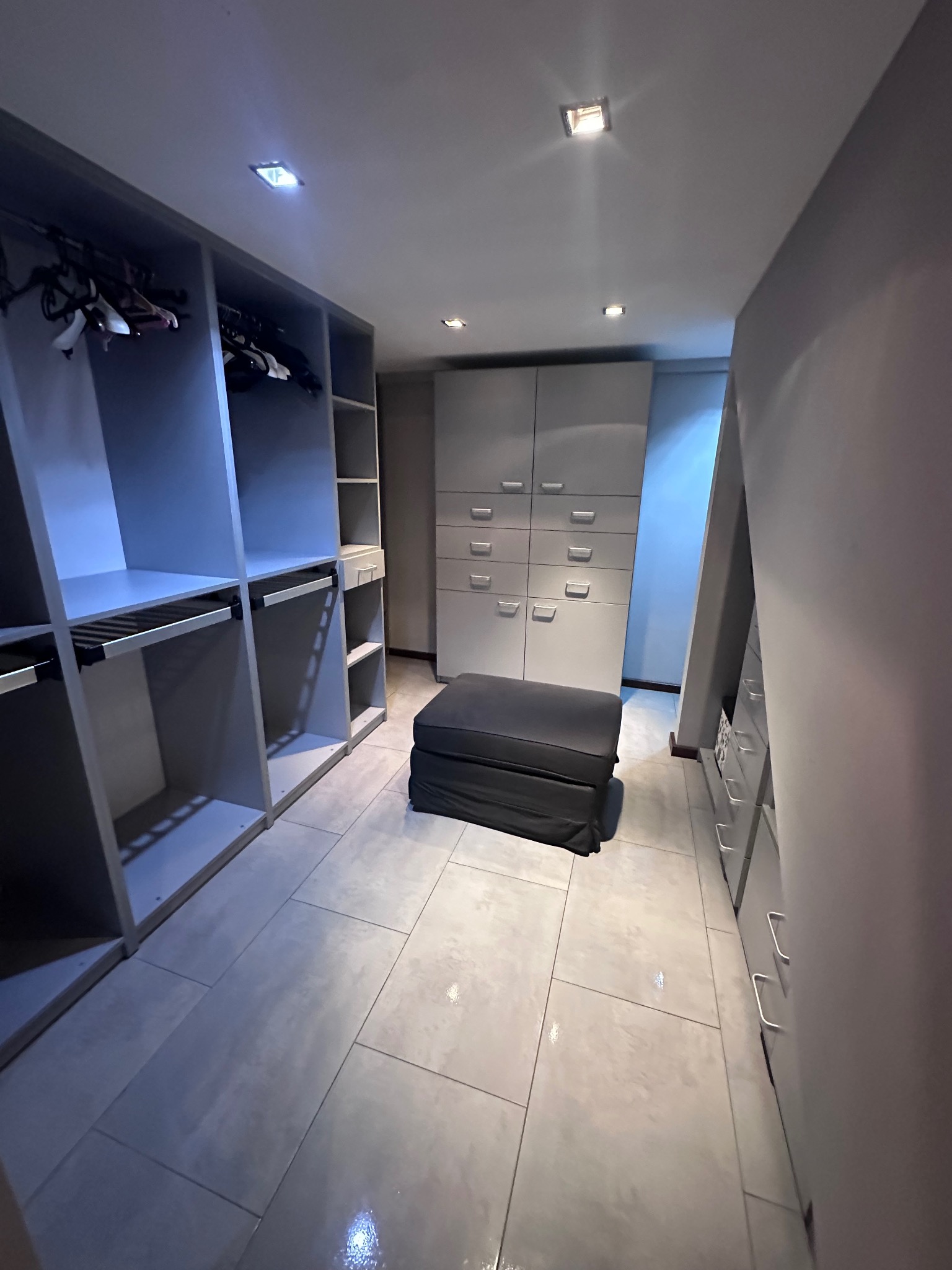 Nowoczesna garderoba z szarymi szafami, półkami i pufą. Minimalistyczny design, jasne płytki podłogowe i punktowe oświetlenie. Widok na uporządkowaną przestrzeń.