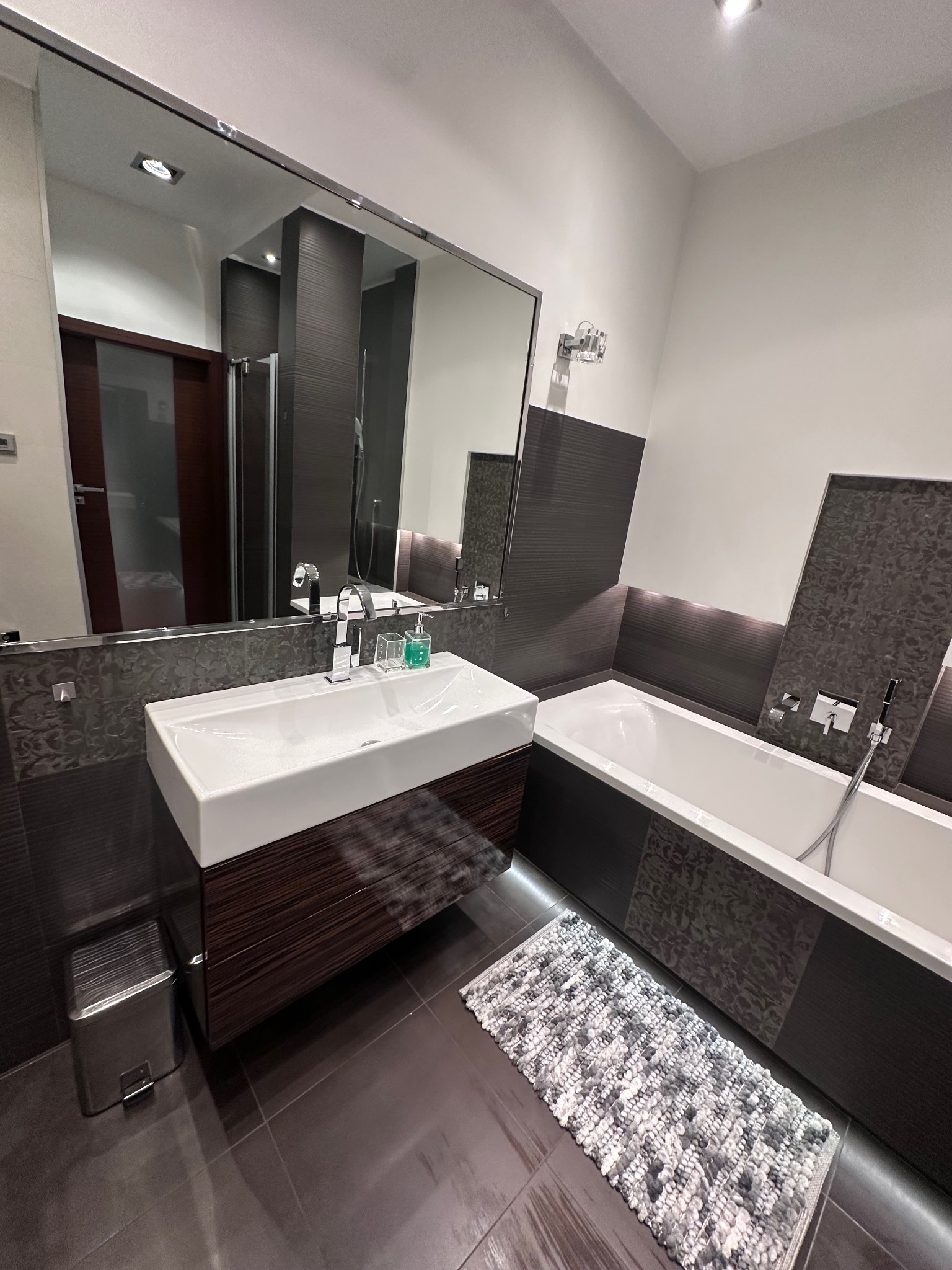 Nowoczesna łazienka z białą wanną, umywalką na szafce z ciemnego drewna i szarą mozaiką. Lustro odbija drzwi i kabinę prysznicową. Ciemna podłoga i dywanik.