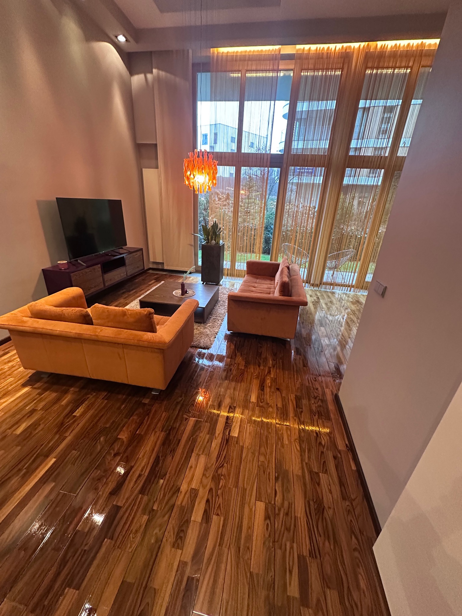 Salon z parkietem w jodełkę, pomarańczowymi sofami i wysokim oknem z zasłonami. Lampa wisząca dodaje ciepła. Widok na zieleń za oknem.