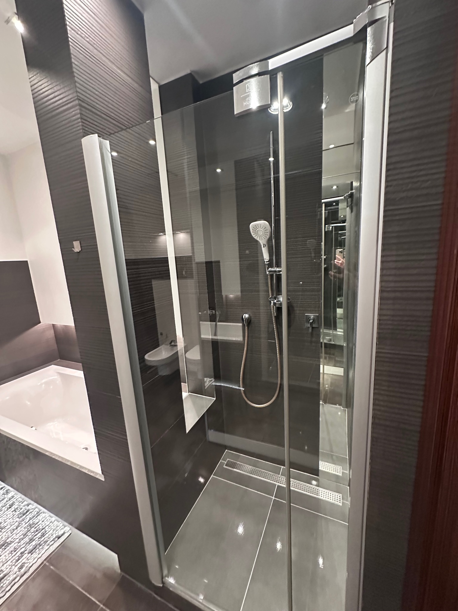 Nowoczesna kabina prysznicowa z grafitowymi płytkami i odpływem liniowym. Widoczna wanna i umywalka w tle. Minimalistyczny design łazienki.