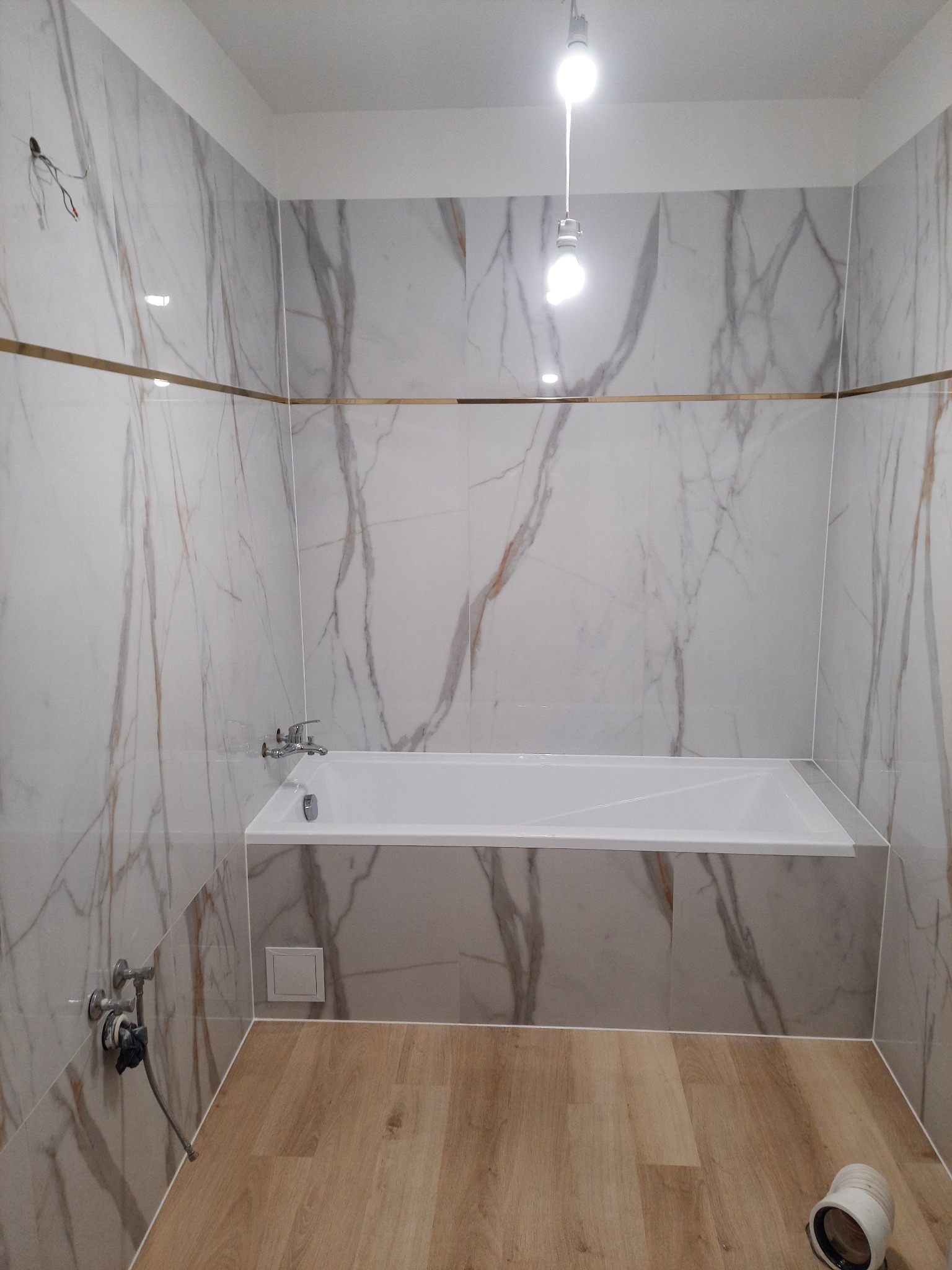 Wykończona łazienka z wanną obudowaną płytkami imitującymi marmur, drewnopodobna podłoga, złote akcenty. Widoczne przyłącza wodne i oświetlenie.