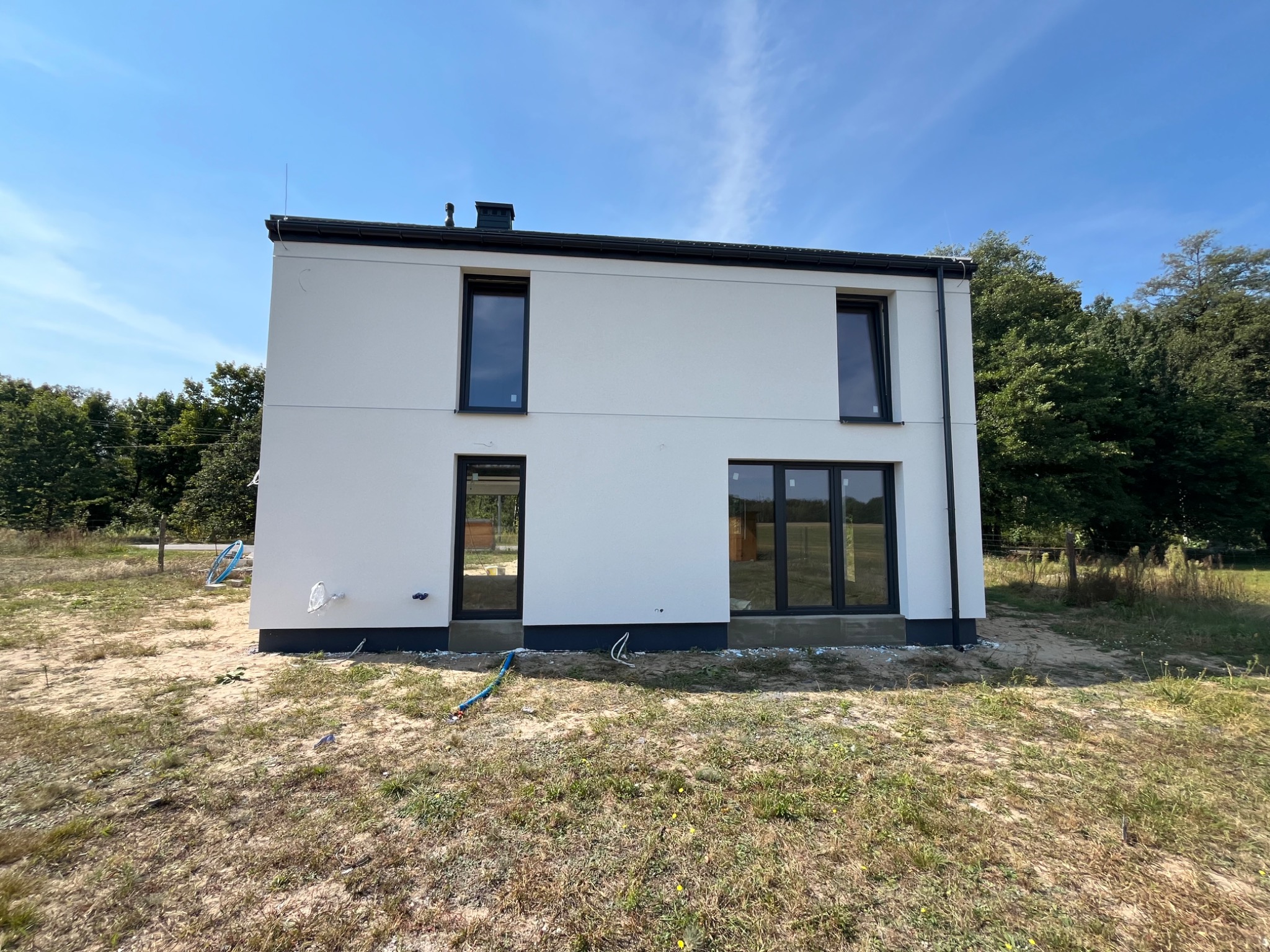 Biały, dwukondygnacyjny dom z czarnymi ramami okiennymi i drzwiami, otoczony trawnikiem i drzewami w słoneczny dzień. Widoczne elementy wykończenia elewacji.