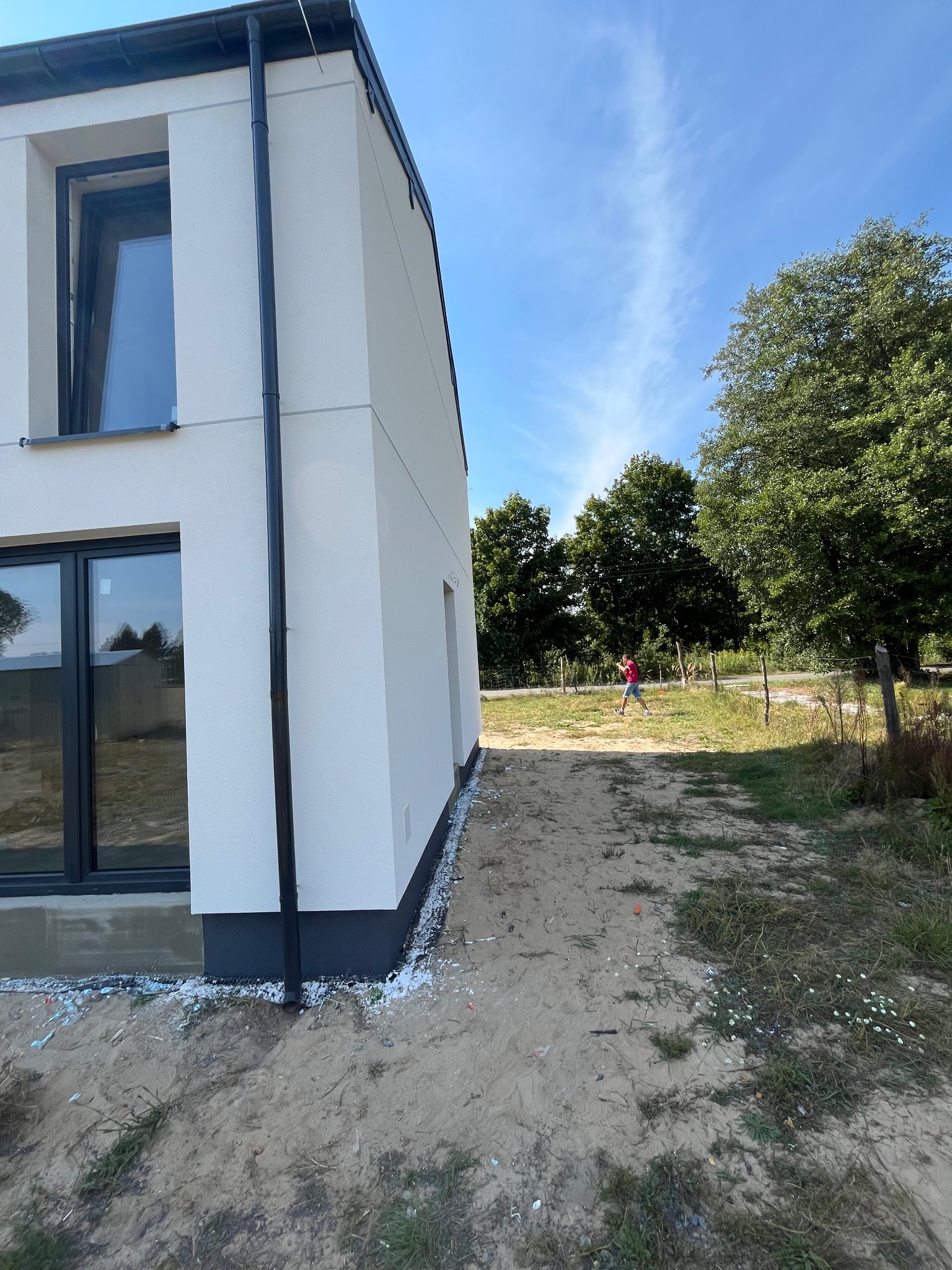 Nowoczesny dom z białą elewacją i czarnymi oknami, widok z boku na tle zieleni. Osoba w oddali. Detale wykończenia elewacji widoczne.