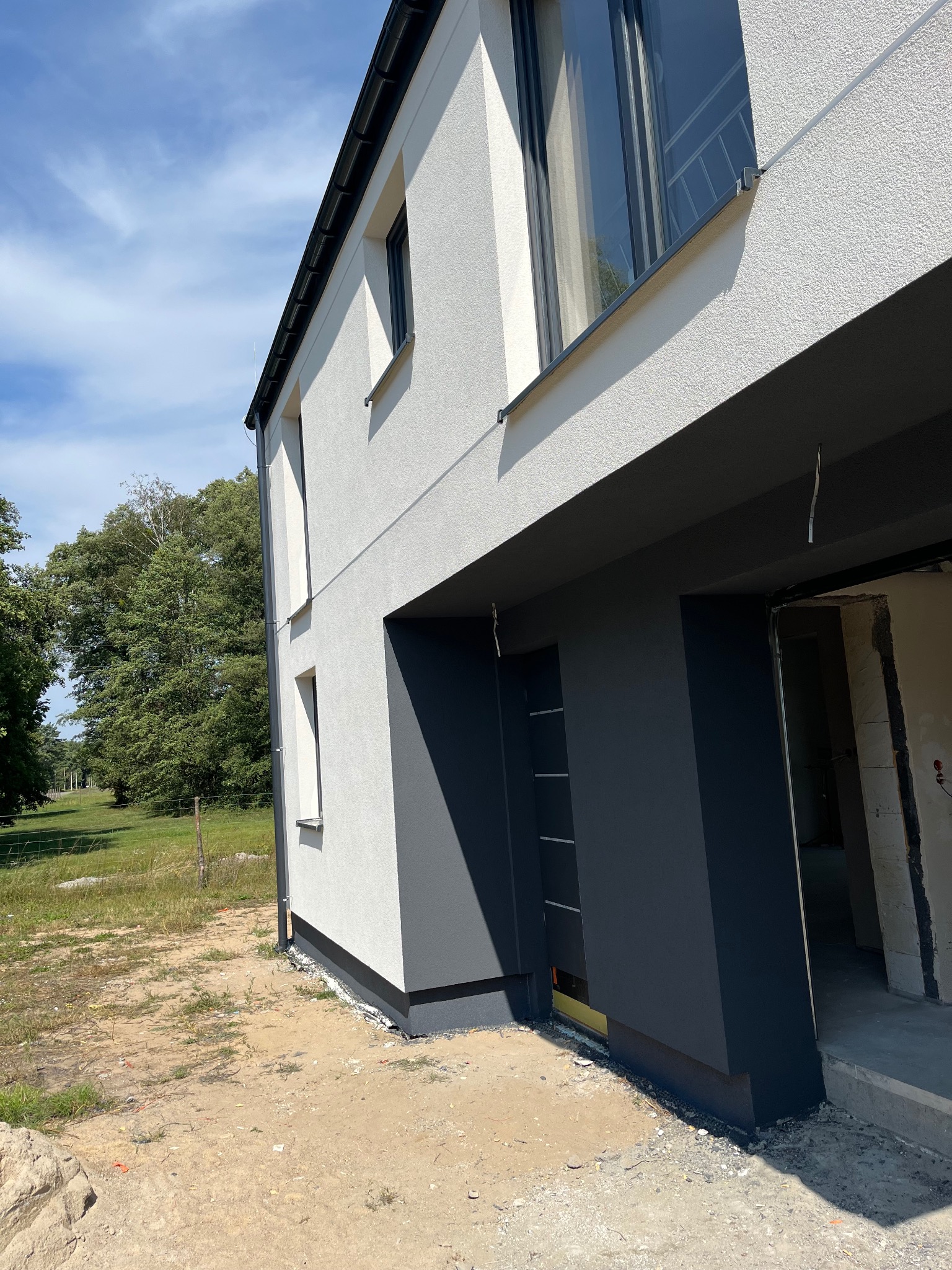 Nowoczesny dom z białą elewacją i grafitowymi akcentami, widok z boku, z fragmentem niezakończonego wejścia. W tle zieleń drzew i błękitne niebo.