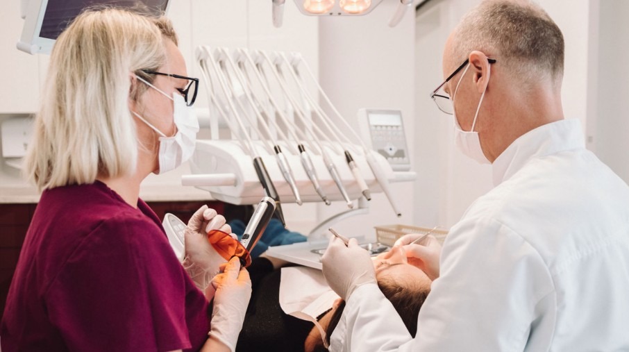Lekarz stomatolog w masce i okularach, asystentka z lampą polimeryzacyjną, pacjent na fotelu dentystycznym, narzędzia stomatologiczne w tle.