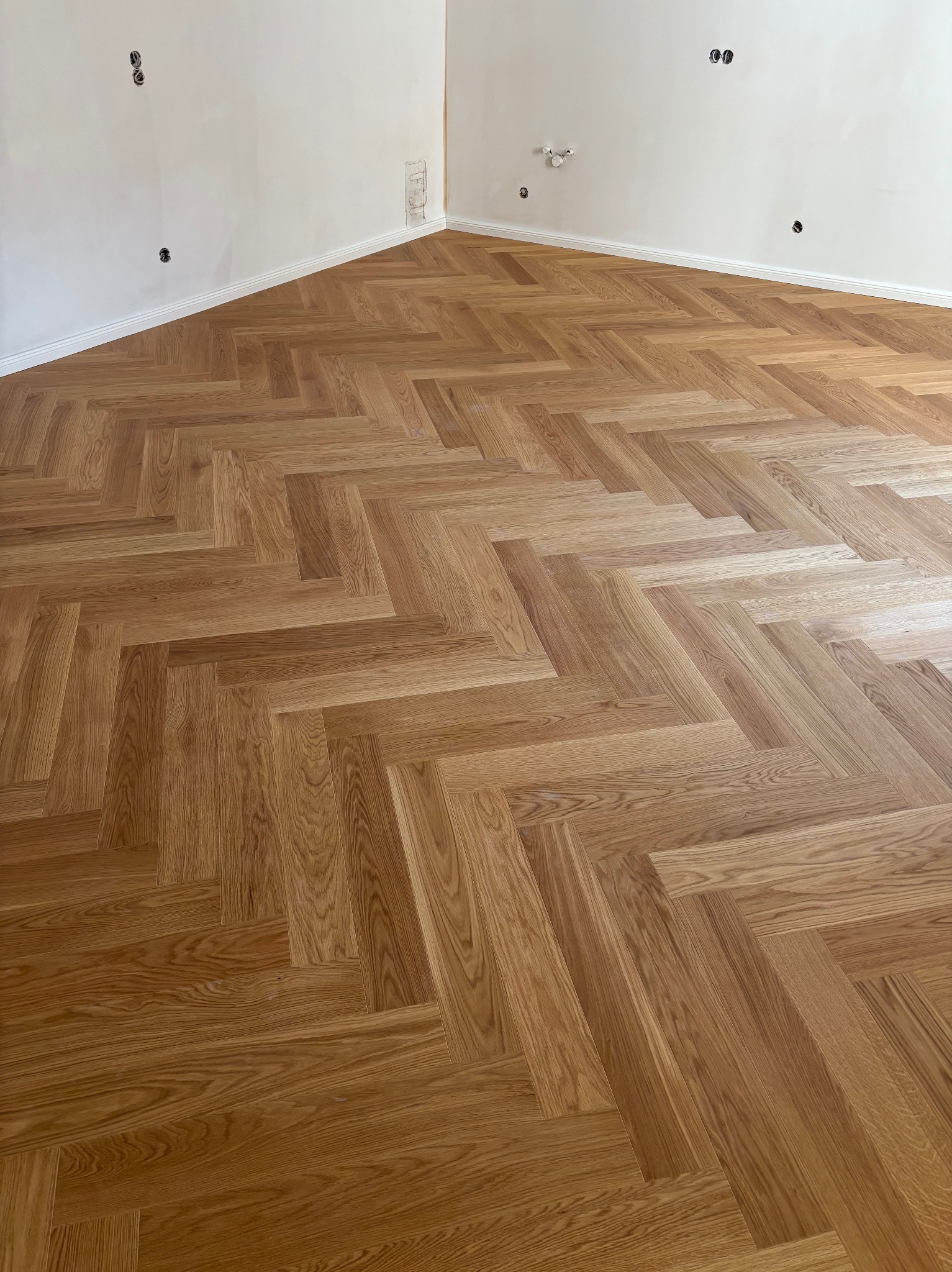 Ukończona podłoga z parkietu ułożonego w jodełkę francuską, widoczne gniazdka elektryczne na ścianie. Jasne drewno, naturalne oświetlenie.