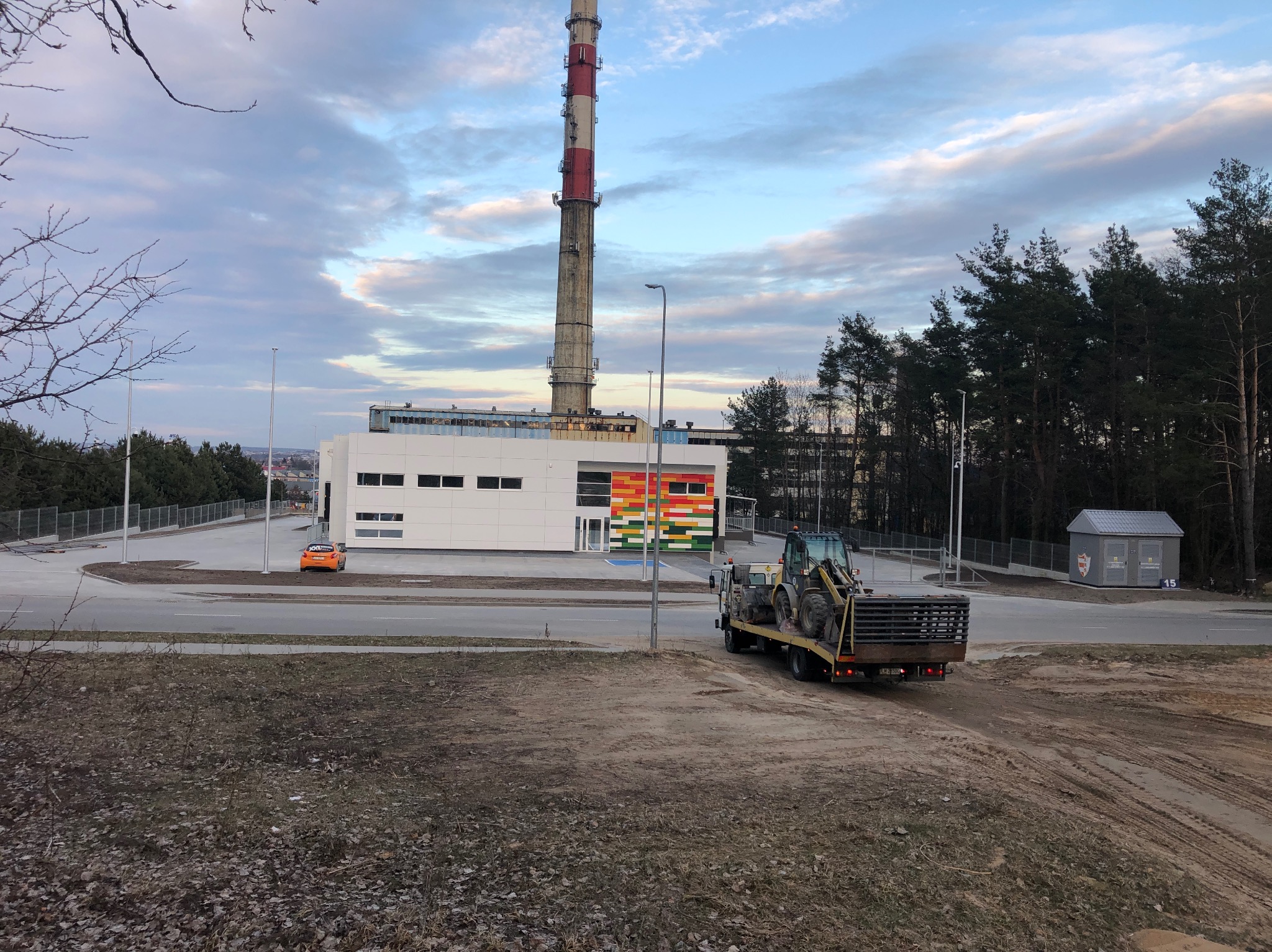 Transport koparki na lawecie w otoczeniu przemysłowym z kominem i budynkiem w tle. Teren przygotowany pod inwestycję, widoczne ślady prac ziemnych.
