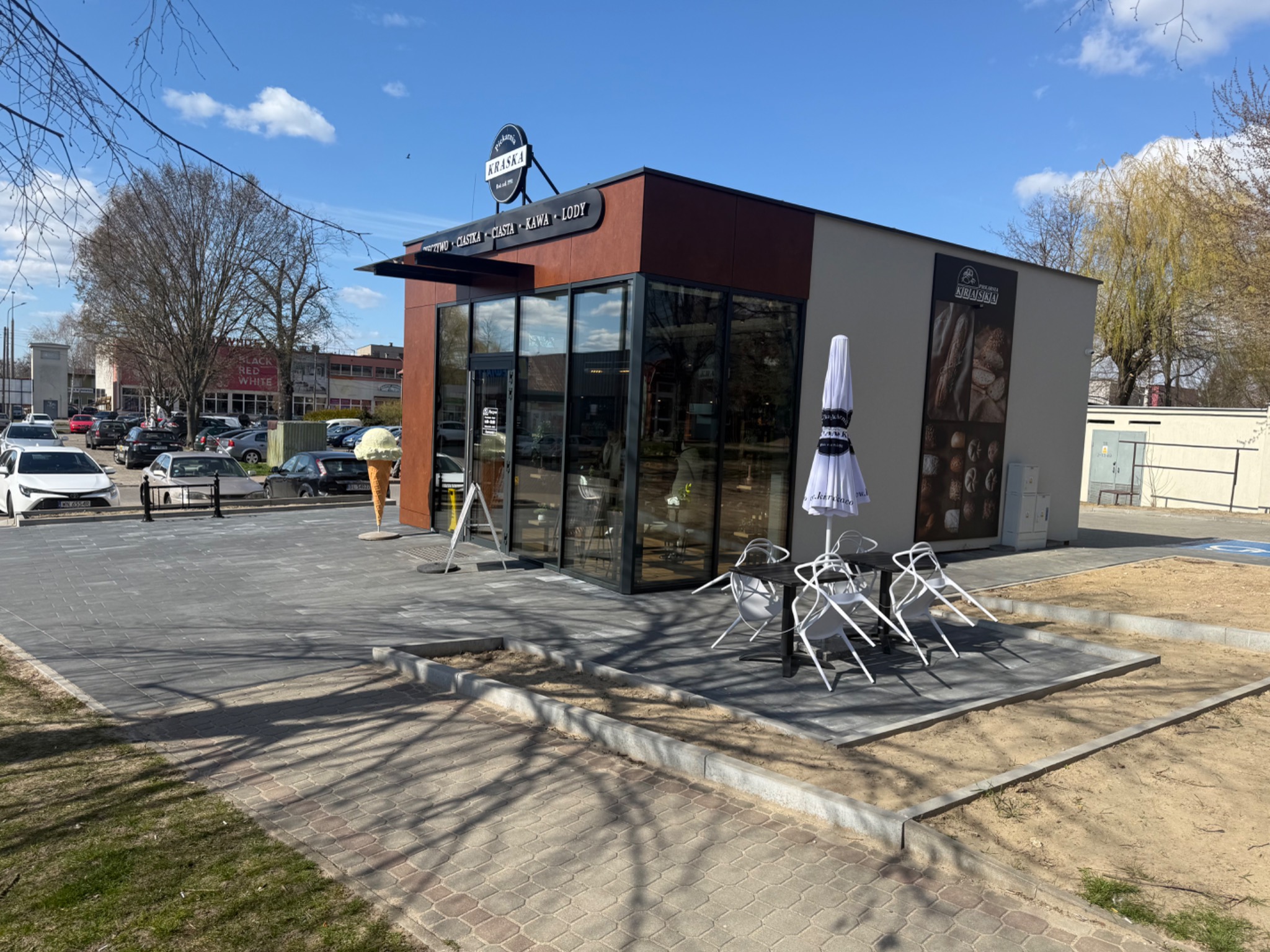 Nowoczesna kawiarnia z kostki brukowej, stoliki na zewnątrz, duży model lodów. Jasne niebo, drzewa w tle. Widoczny parking i inne budynki. Słoneczny dzień.